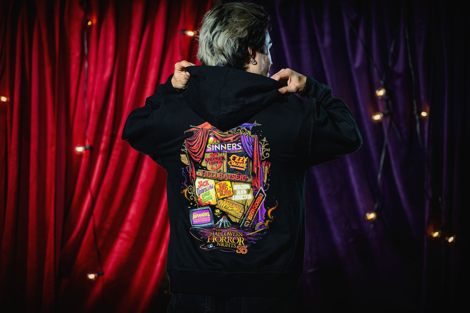 A person seen from behind pulling up the hood of a black sweatshirt whose back print stacks the names of every Halloween Horror Nights 35 house above the event logo