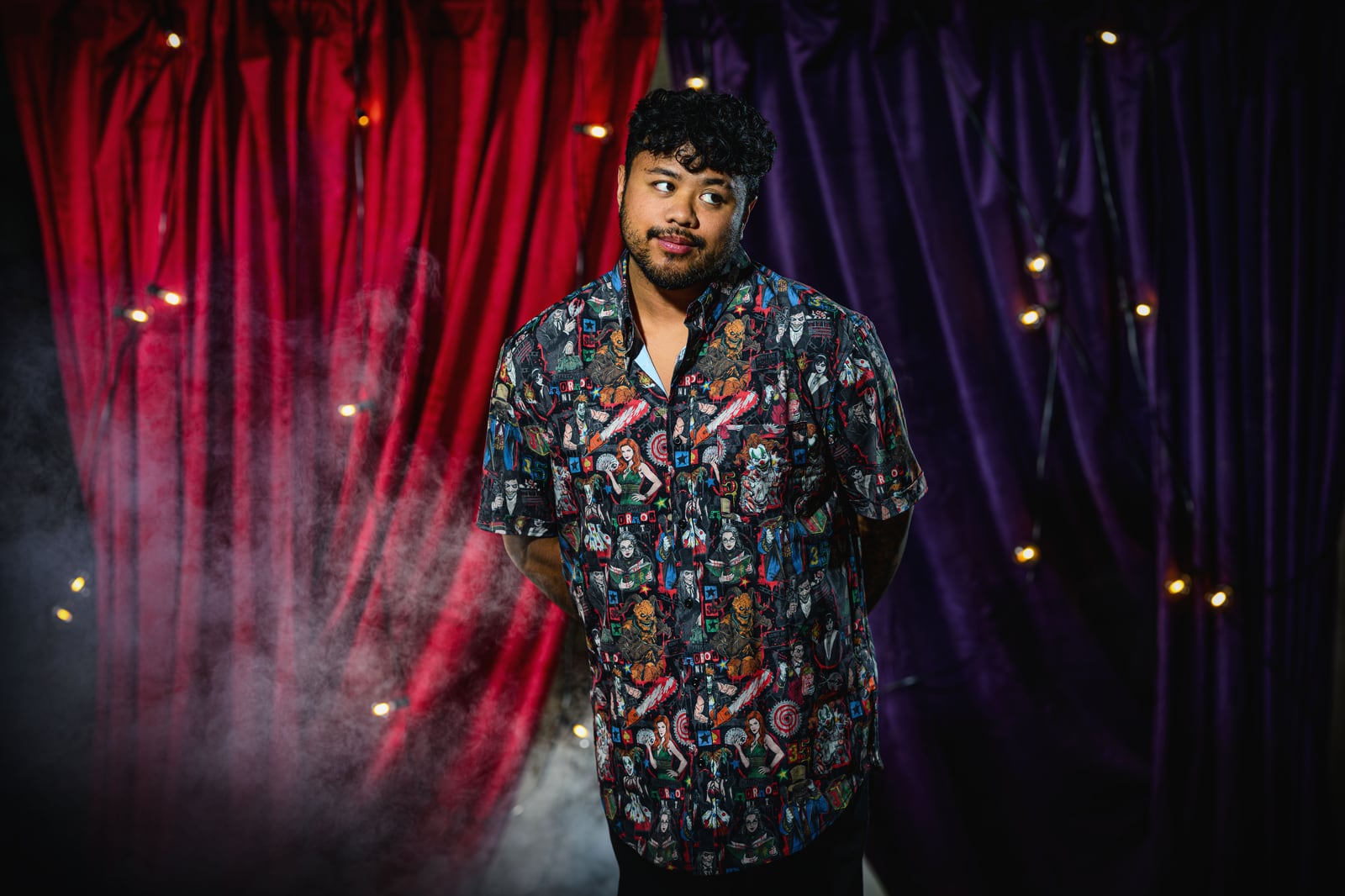 A man wearing a short-sleeved button-up shirt printed edge to edge with Halloween Horror Nights icon artwork, standing in front of red and purple curtains with fog