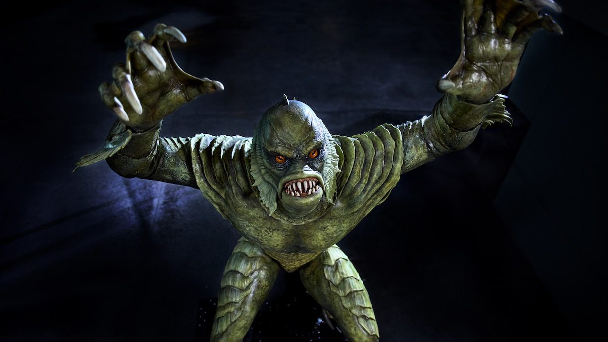 The Creature from the Black Lagoon figure from Monsters Unchained at Epic Universe, a green scaled humanoid with orange eyes and both clawed hands raised, lit against a dark background.