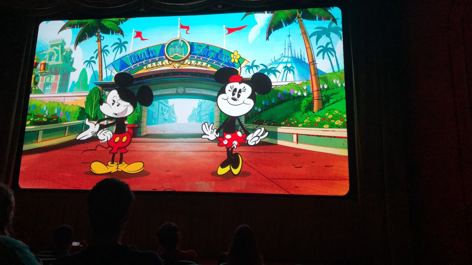 Mickey and Minnie Mouse animated on the Mickey Shorts Theater screen, standing in front of a park main entrance archway, with silhouettes of the audience in the foreground