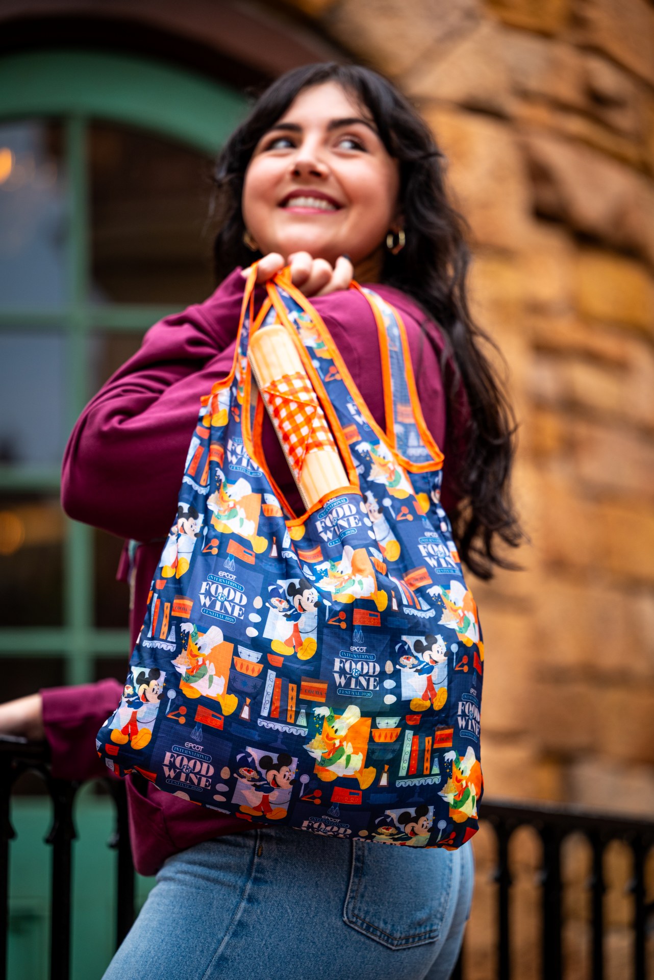 A guest carries the 2026 EPCOT Food and Wine Festival tote bag over one shoulder, printed all over with Mickey and friends cooking, hanging from orange straps with a ridged tan churro shaped pouch attached to them