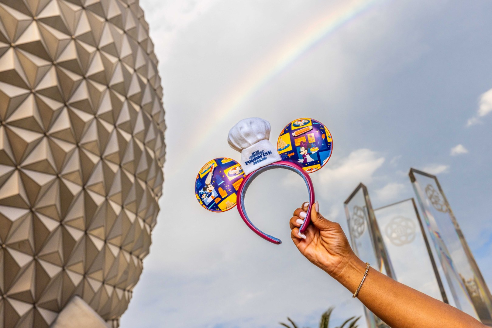 A hand holds up the 2026 EPCOT Food and Wine Festival Mickey ears, topped with a white chef hat and printed with colorful food panels, in front of Spaceship Earth with a rainbow overhead