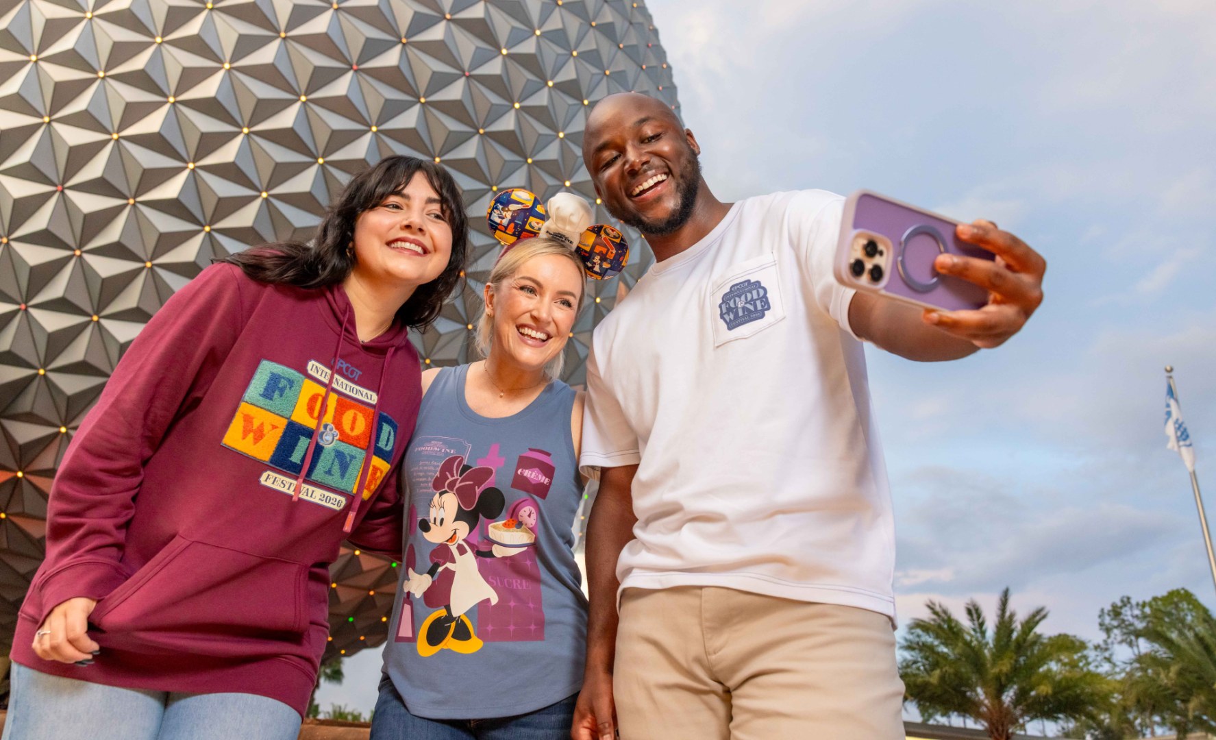 Three guests take a selfie in front of Spaceship Earth wearing 2026 EPCOT Food and Wine Festival merchandise, including a deep red hooded sweatshirt with a colorful block letter patch, a blue Minnie Mouse chef tank top, and a white pocket tee