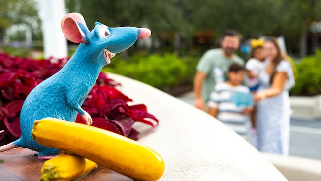A small blue Remy statuette from Ratatouille stands on a ledge beside a yellow squash at EPCOT, with a family blurred in the background