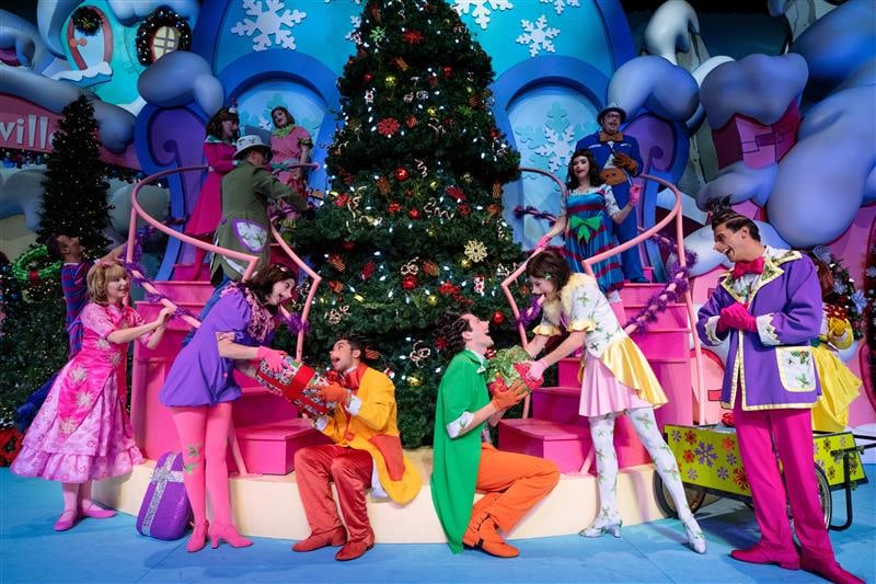 Performers in bright Who-ville costumes dance around a decorated Christmas tree on the Grinchmas stage set.