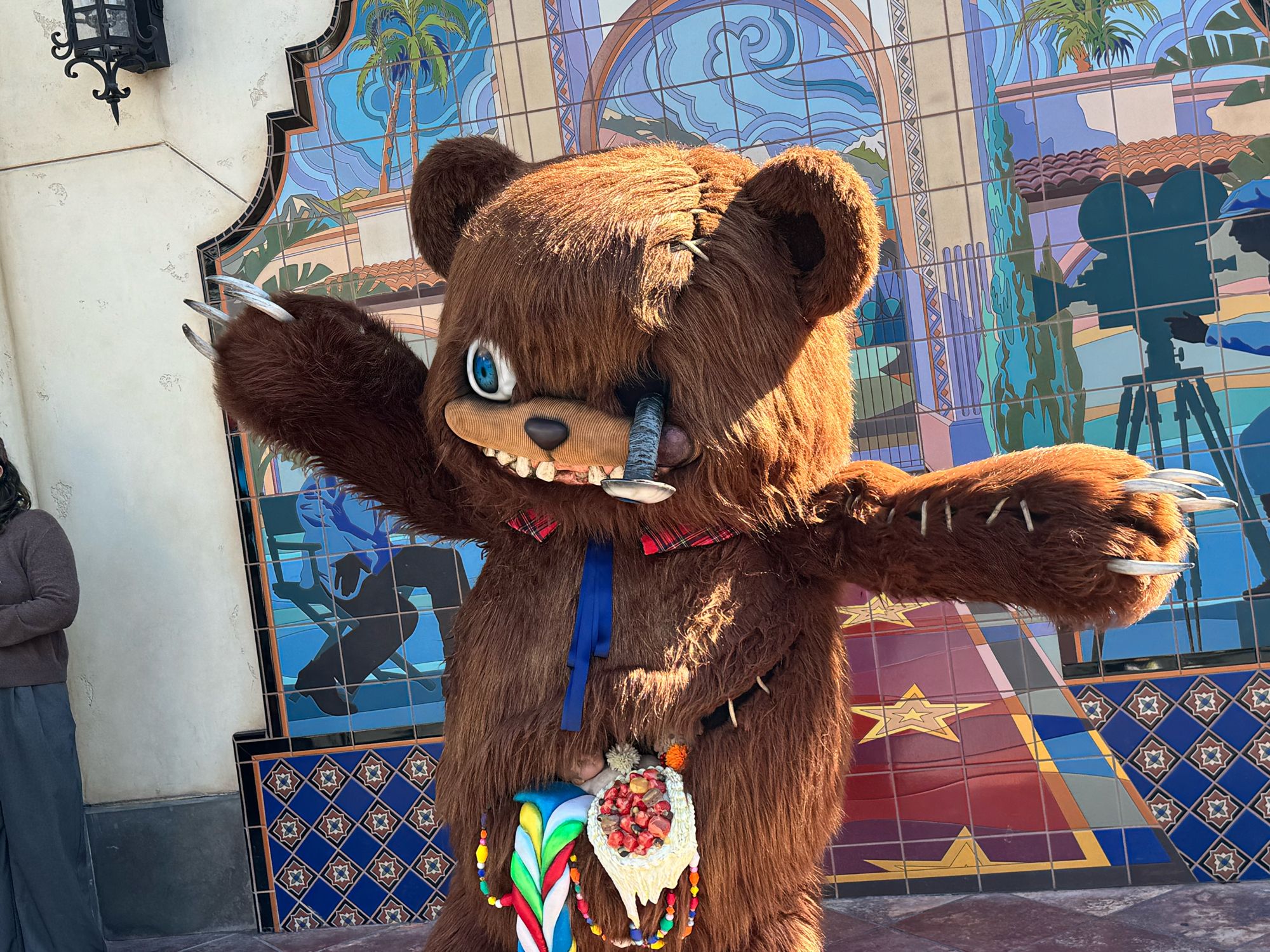 HamiKuma in a full walkaround costume with both arms stretched wide and long claws extended, standing in front of a tiled mural of palm trees and a film camera