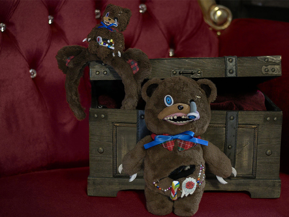 Two HamiKuma plush toys with blue bow ties and candy spilling from their open bellies, one draped over the lid of an open wooden chest and one seated in front of it against a red tufted velvet background