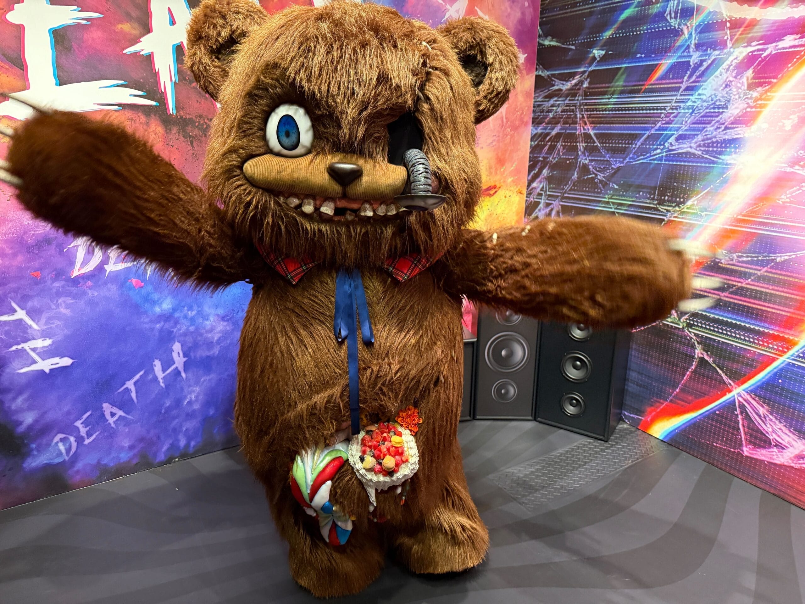 HamiKuma in costume with arms outstretched inside a meet and greet set at Universal Studios Japan, his loose eye dangling on its stalk over his teeth, standing in front of a neon pink and purple glitch art backdrop with stereo speakers