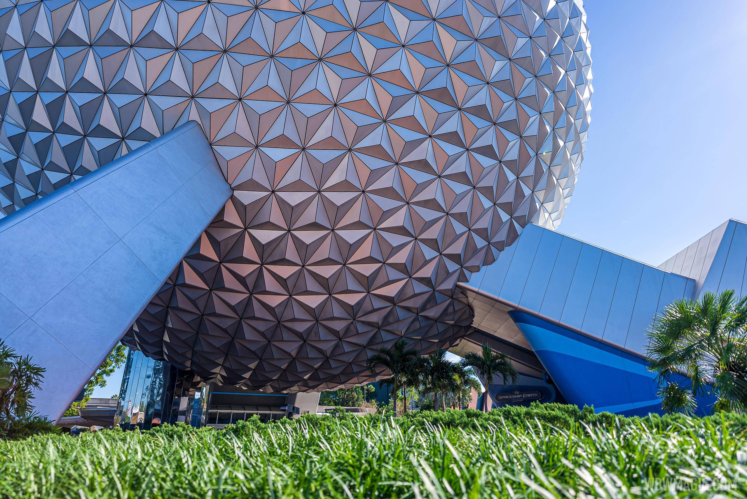 A low angle view of the underside of Spaceship Earth at EPCOT, showing silver triangular panels curving overhead, the angled legs supporting the sphere, and a Spaceship Earth sign at right.