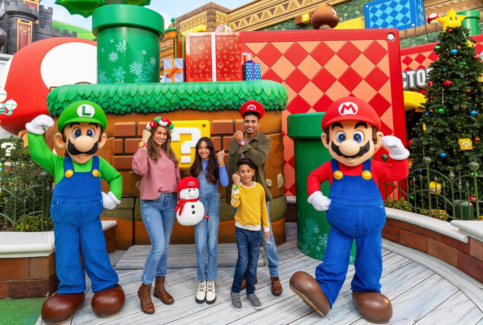 Mario and Luigi pose with a family in Super Nintendo World, with wrapped gift boxes stacked on a warp pipe, snowflake decals on the green pipes, and a decorated Christmas tree at right.
