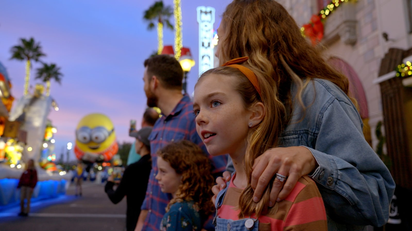 A family watches Universal's Holiday Parade featuring Macy's at dusk, with a giant Minion balloon floating down the street behind them.
