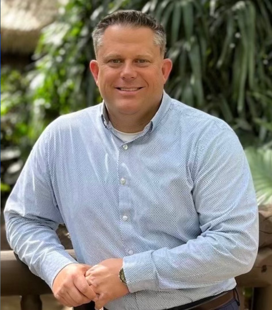 Portrait of Brad Gilmour, smiling in a light blue button-down shirt, standing in front of tropical foliage.