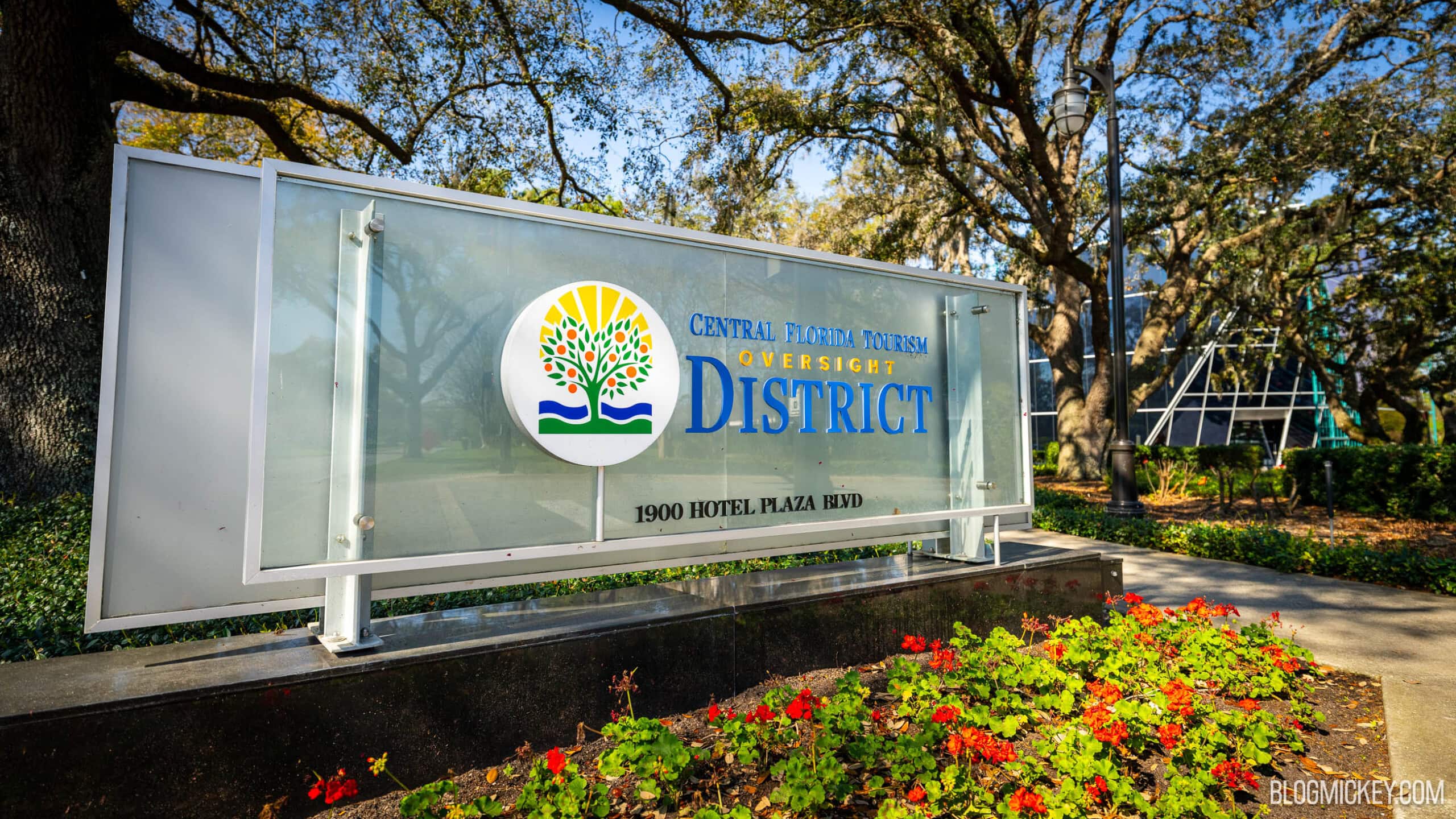 A glass and metal monument sign reading Central Florida Tourism Oversight District, with the district's orange tree and sunrise logo, standing in front of live oak trees and a bed of red flowers.
