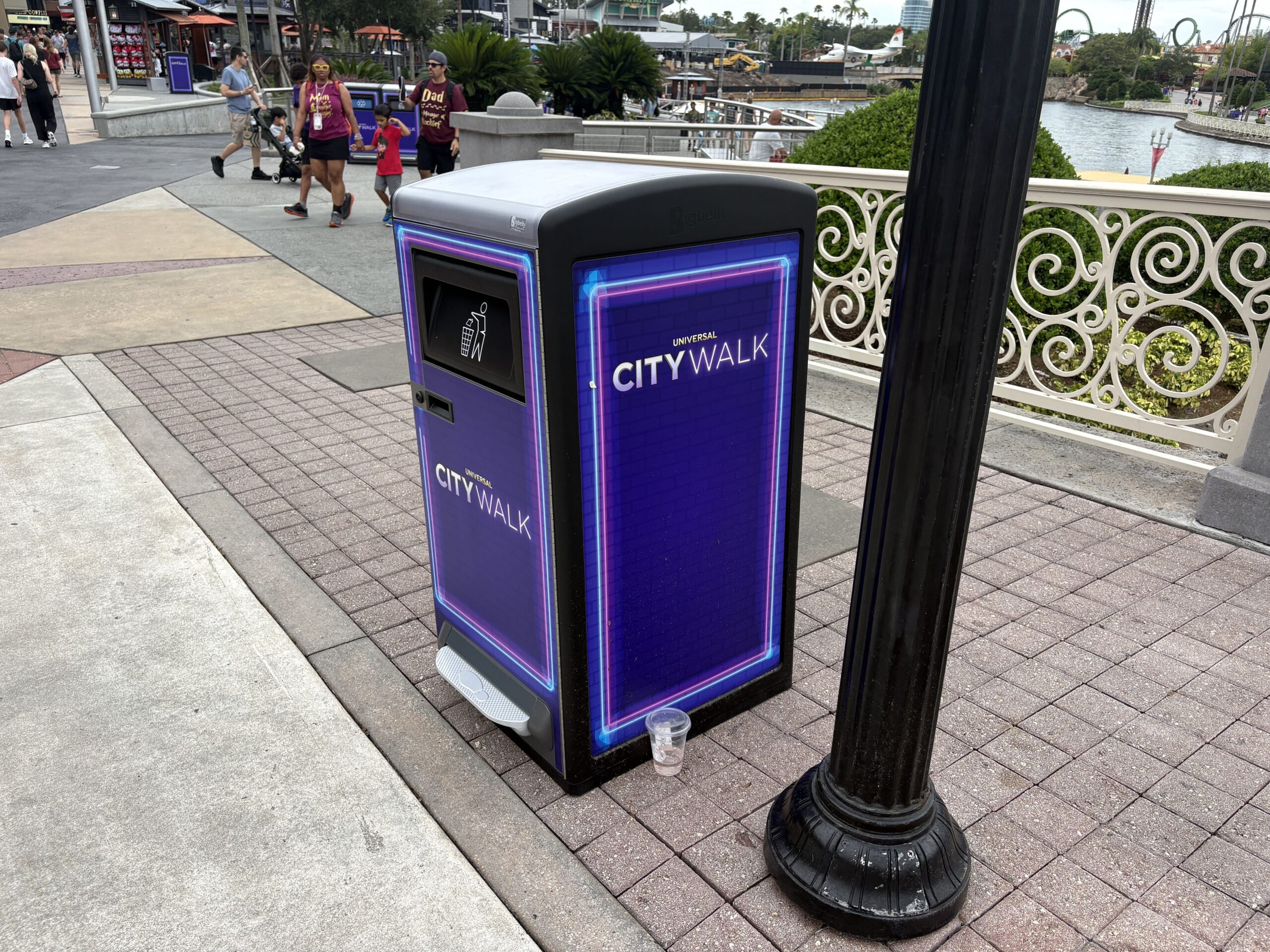 A single purple neon trash bin stands on a brick walkway next to a black lamppost, with a clear plastic cup left on the ground beside its base and guests walking along CityWalk in the background.