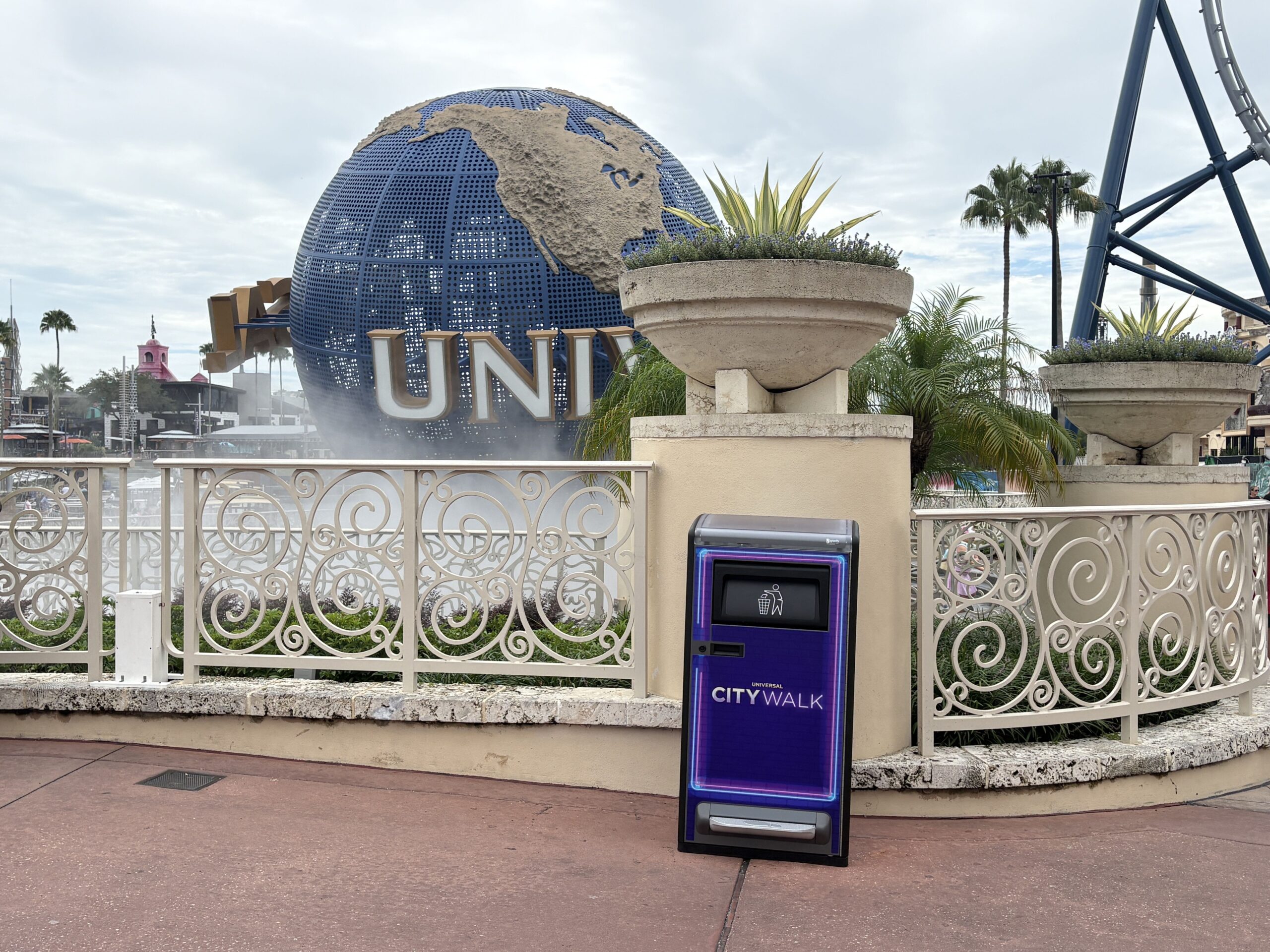 A tall purple trash bin with neon blue and pink outlines and a Universal CityWalk logo stands on a walkway in front of the rotating Universal globe fountain, with palm trees and a roller coaster track behind it.