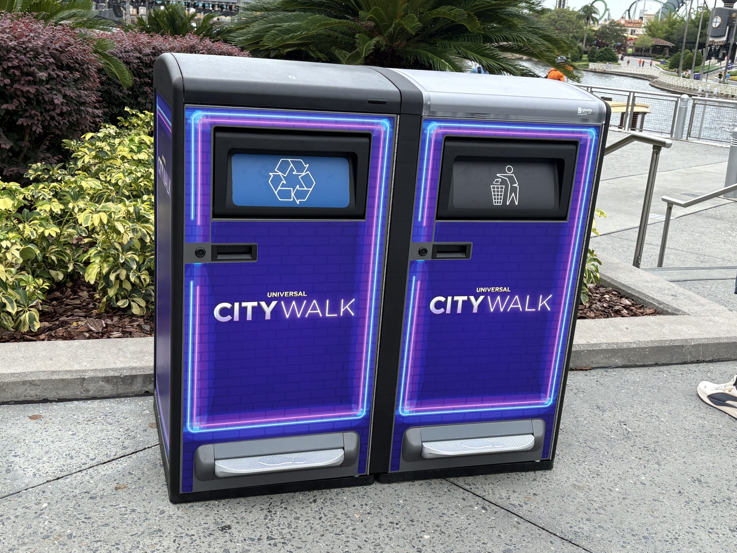 A paired purple recycling bin and trash bin with neon outlines and Universal CityWalk logos, each with a small opening at the top and a metal foot pedal along the bottom, on a walkway beside landscaping.