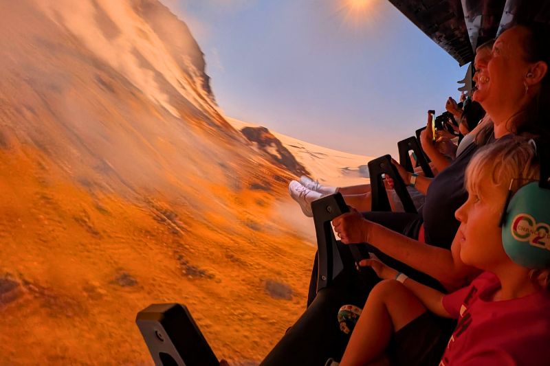 Riders in a SeaWorld Orlando attraction vehicle facing a wraparound screen showing an orange volcanic mountainside, with a child in green ear defenders in the foreground.