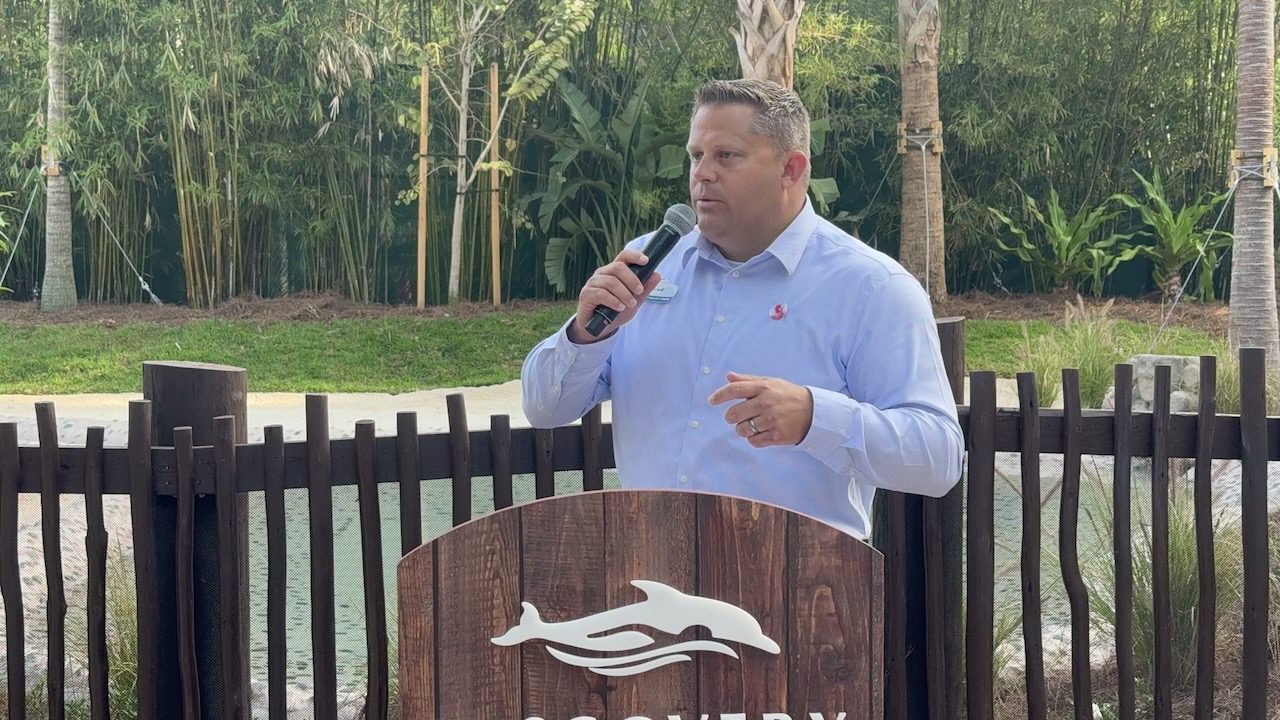 Brad Gilmour speaking into a microphone behind a wooden podium bearing the Discovery Cove dolphin logo, with bamboo and a lagoon behind him.