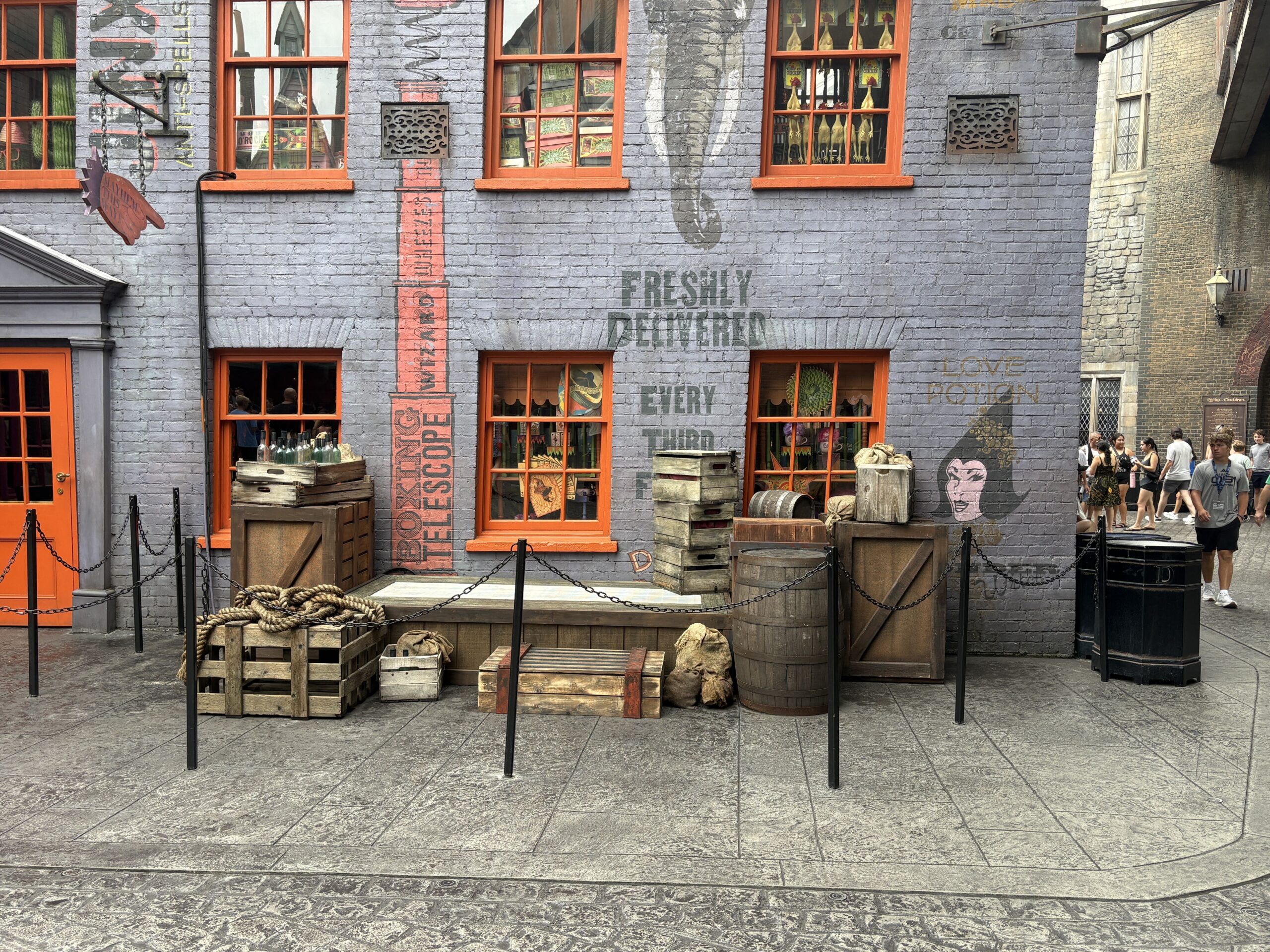 A low wooden platform in Diagon Alley surrounded by stacked crates, barrels, coiled rope and burlap sacks, roped off with chain stanchions against the lavender-grey brick wall behind Weasleys' Wizard Wheezes.
