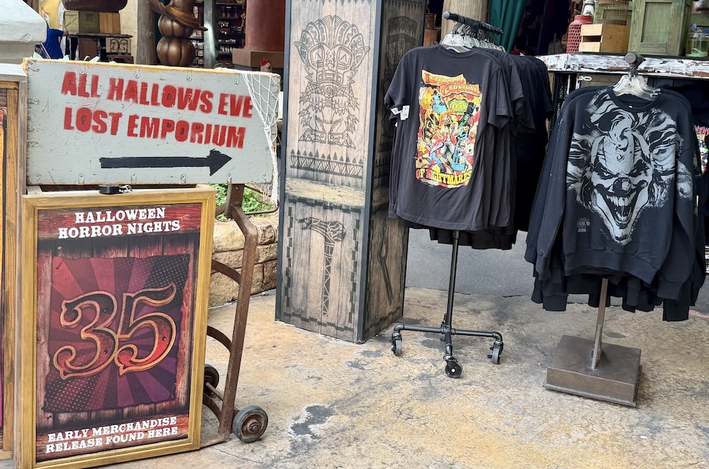 A weathered sign reading All Hallows Eve Lost Emporium with an arrow, above a framed Halloween Horror Nights 35 easel sign reading early merchandise release found here, next to racks of black shirts and a clown-face sweatshirt.