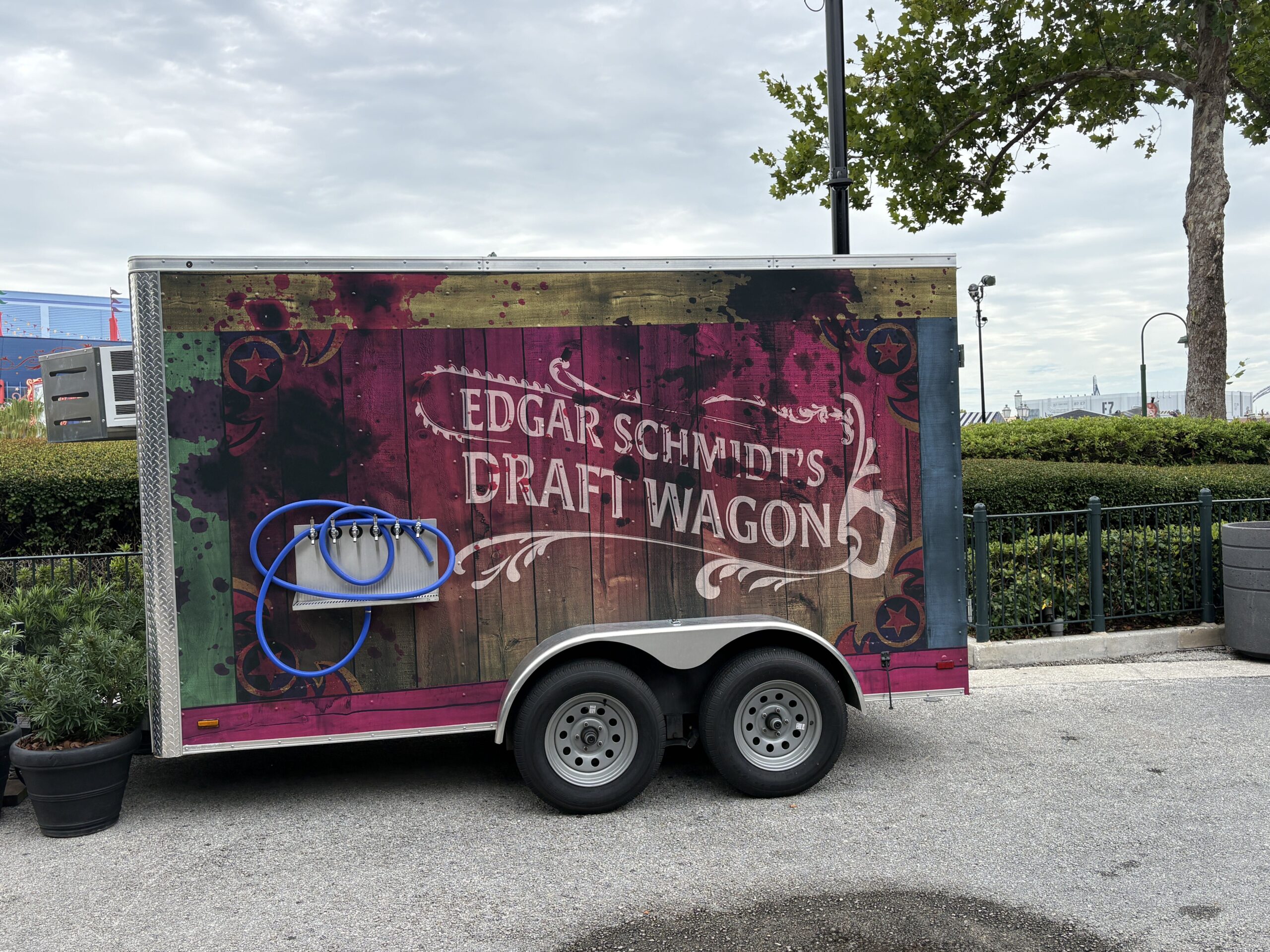 A beverage trailer wrapped in distressed wood, magenta and green artwork splattered with dark blood stains, with a bank of six taps and a coiled blue hose on its side, and lettering reading Edgar Schmidt's Draft Wagon.
