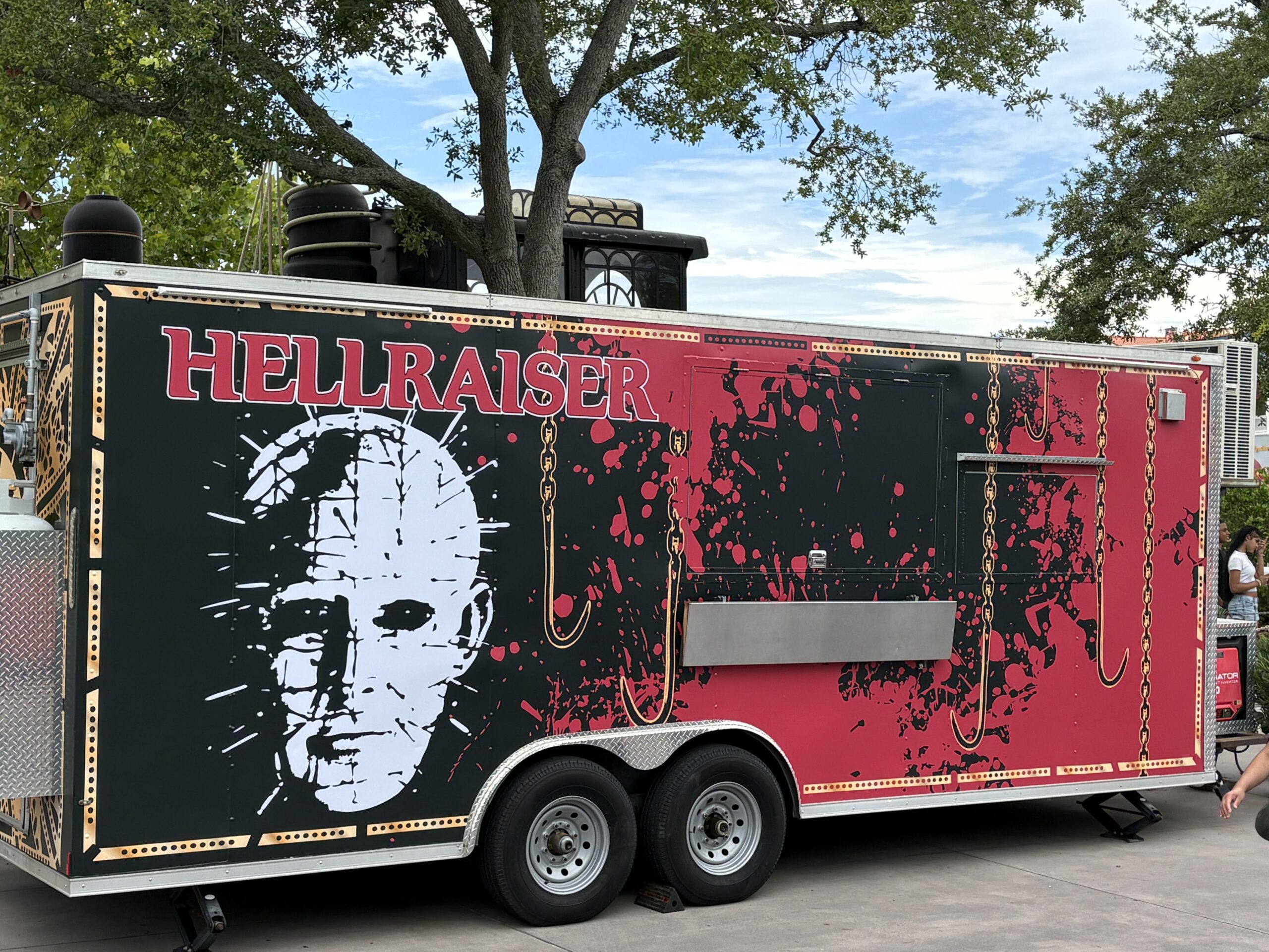 A black and red concession trailer wrapped with the Hellraiser logo and a large white illustration of Pinhead's pinned face, with dangling hook and chain artwork and red splatter across the side panels beside a closed serving window.