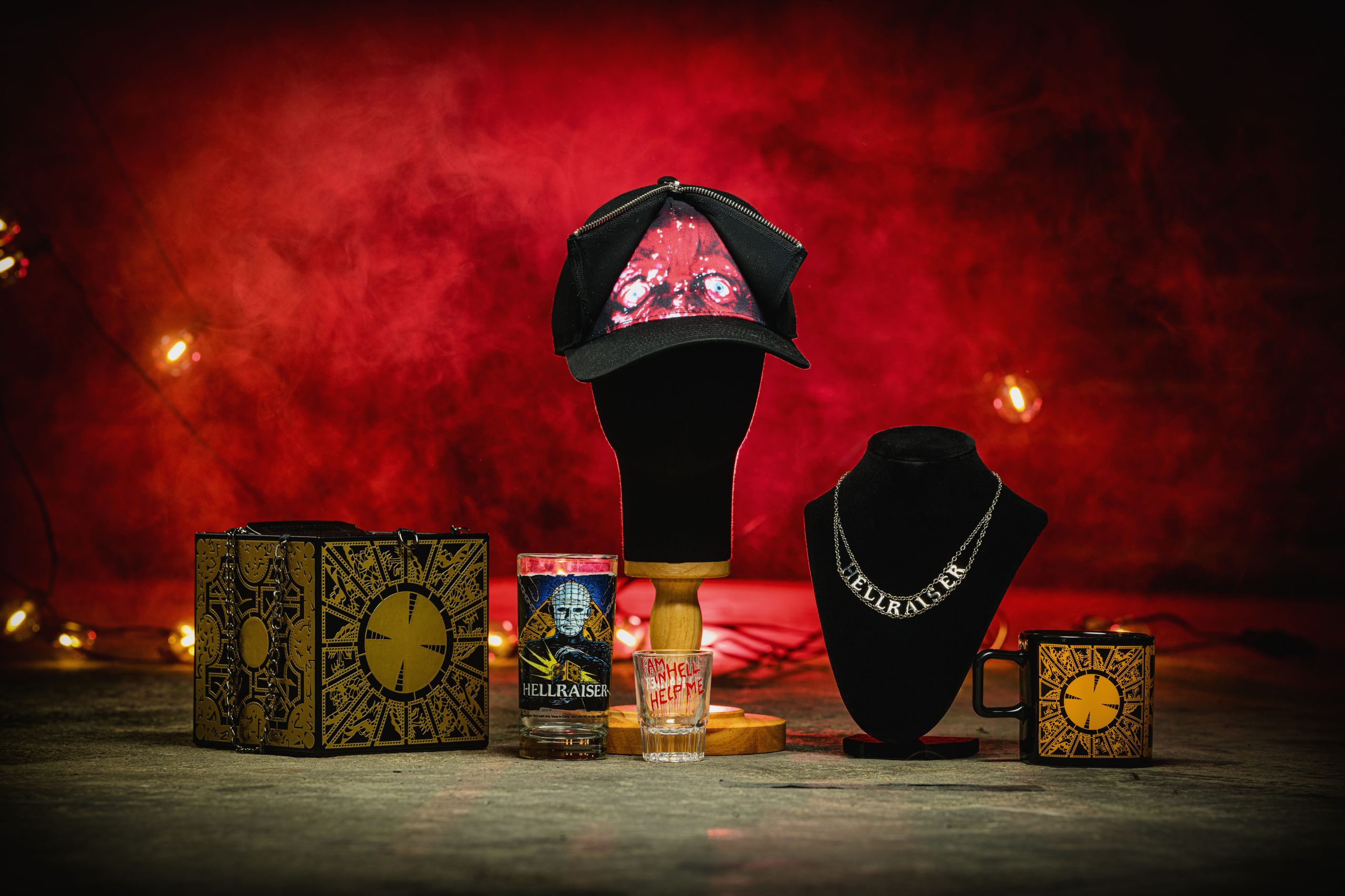 A Hellraiser merchandise display against a red smoky background, showing a gold Lament Configuration puzzle box purse, a Pinhead pint glass, a shot glass reading I am in hell help me, a cap with a Cenobite face print, a chain necklace spelling Hellraiser and a Lament Configuration mug.