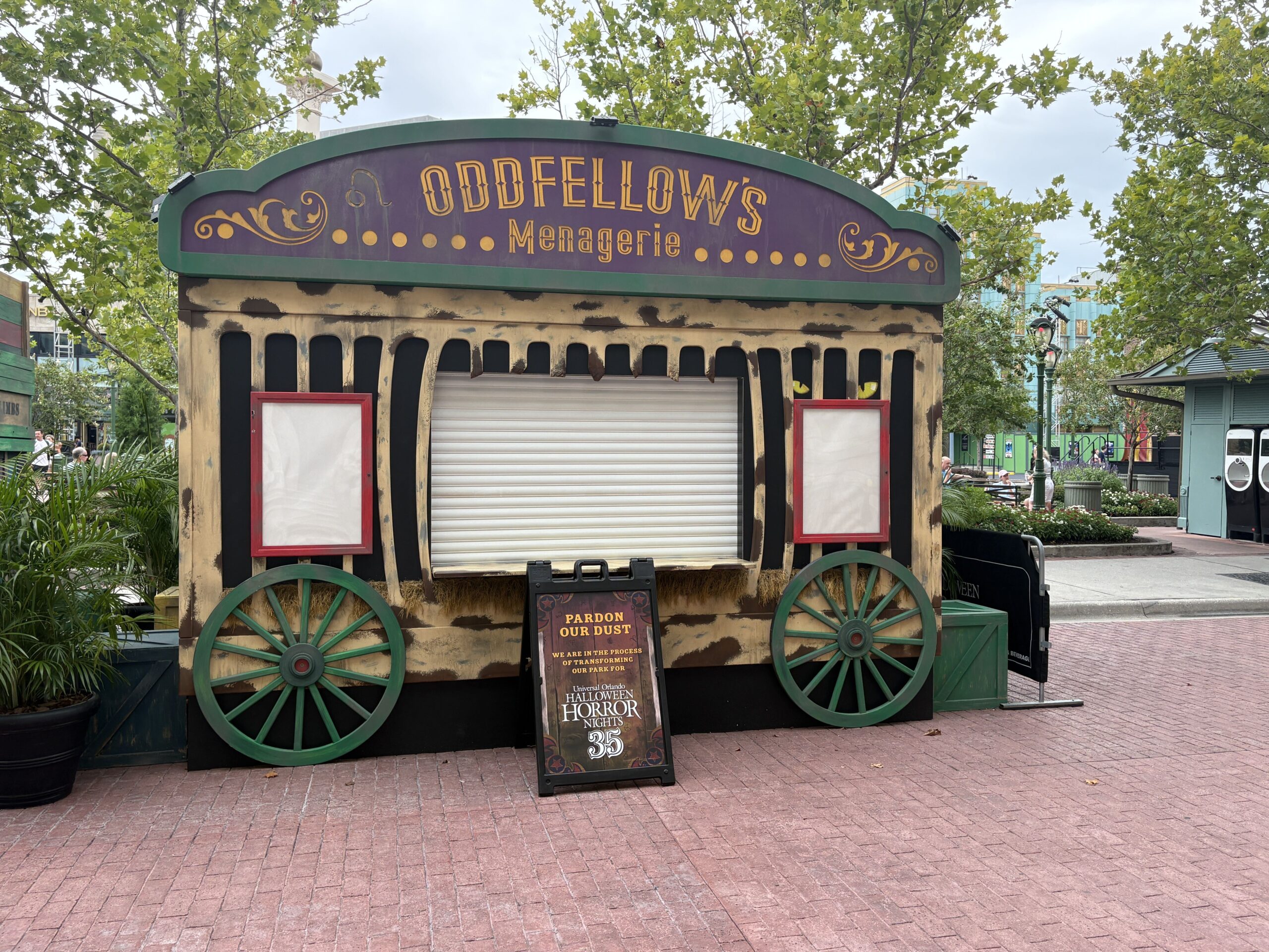 A carnival wagon food booth with green spoked wheels, a distressed cream body and a purple arched sign reading Oddfellow's Menagerie in gold lettering, its serving shutter still closed, with a Pardon Our Dust sandwich board in front.