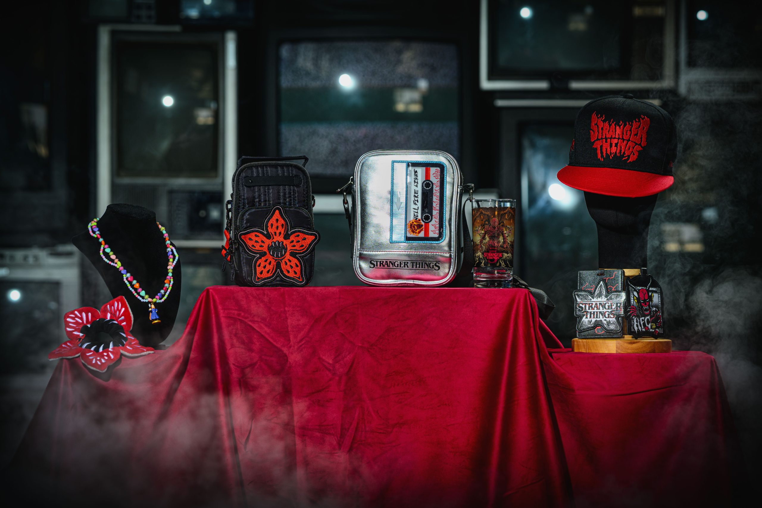 A Stranger Things merchandise display on a red velvet table, including a black bag with an orange Demogorgon patch, a silver cassette-tape shoulder bag, a Vecna pint glass, a Stranger Things cap, a beaded necklace and a Hellfire Club bag clip.