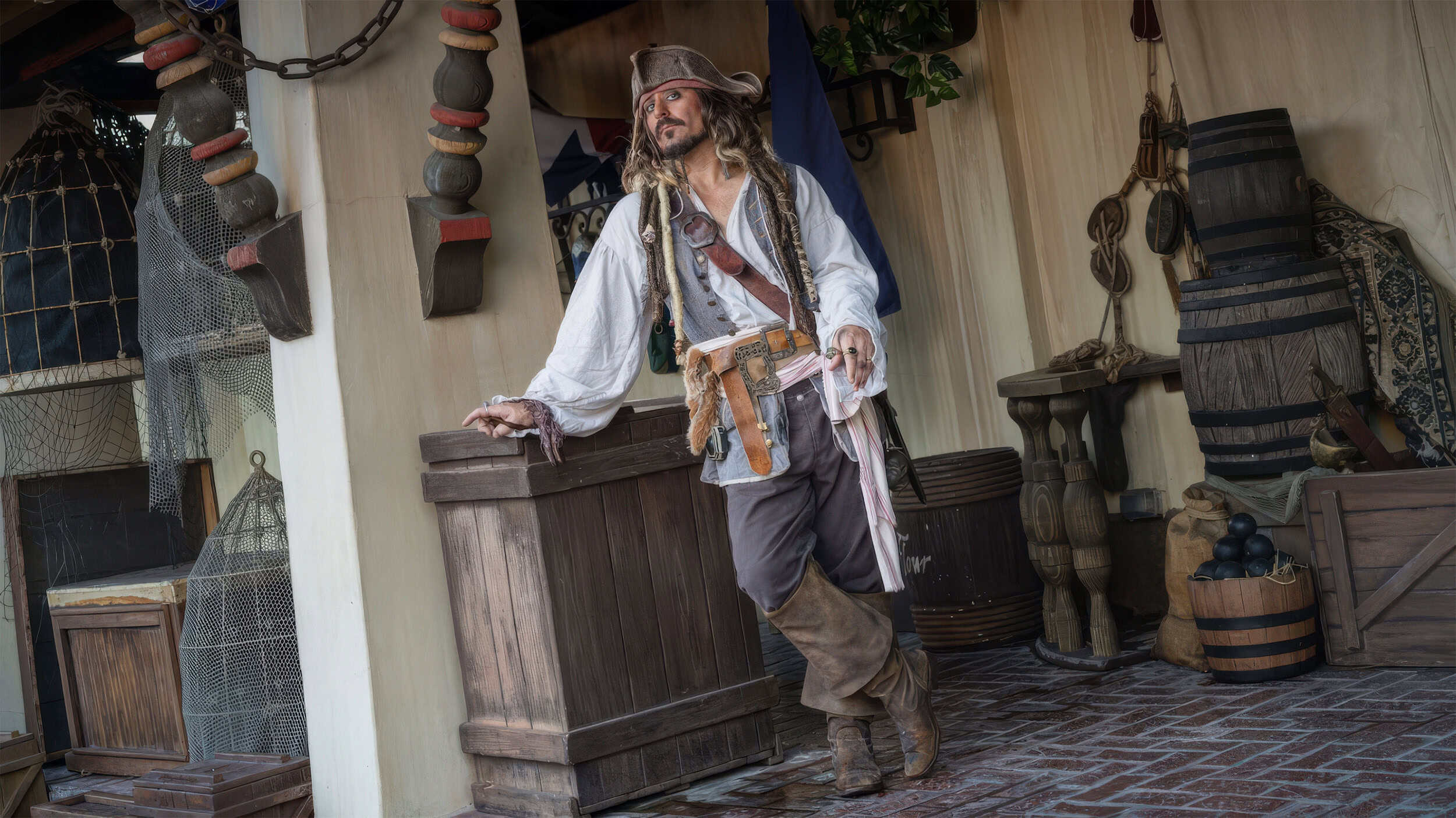 Captain Jack Sparrow leaning against a wooden crate on the pirate-dressed Adventureland set at Magic Kingdom, surrounded by barrels, fishing nets and hanging cages.
