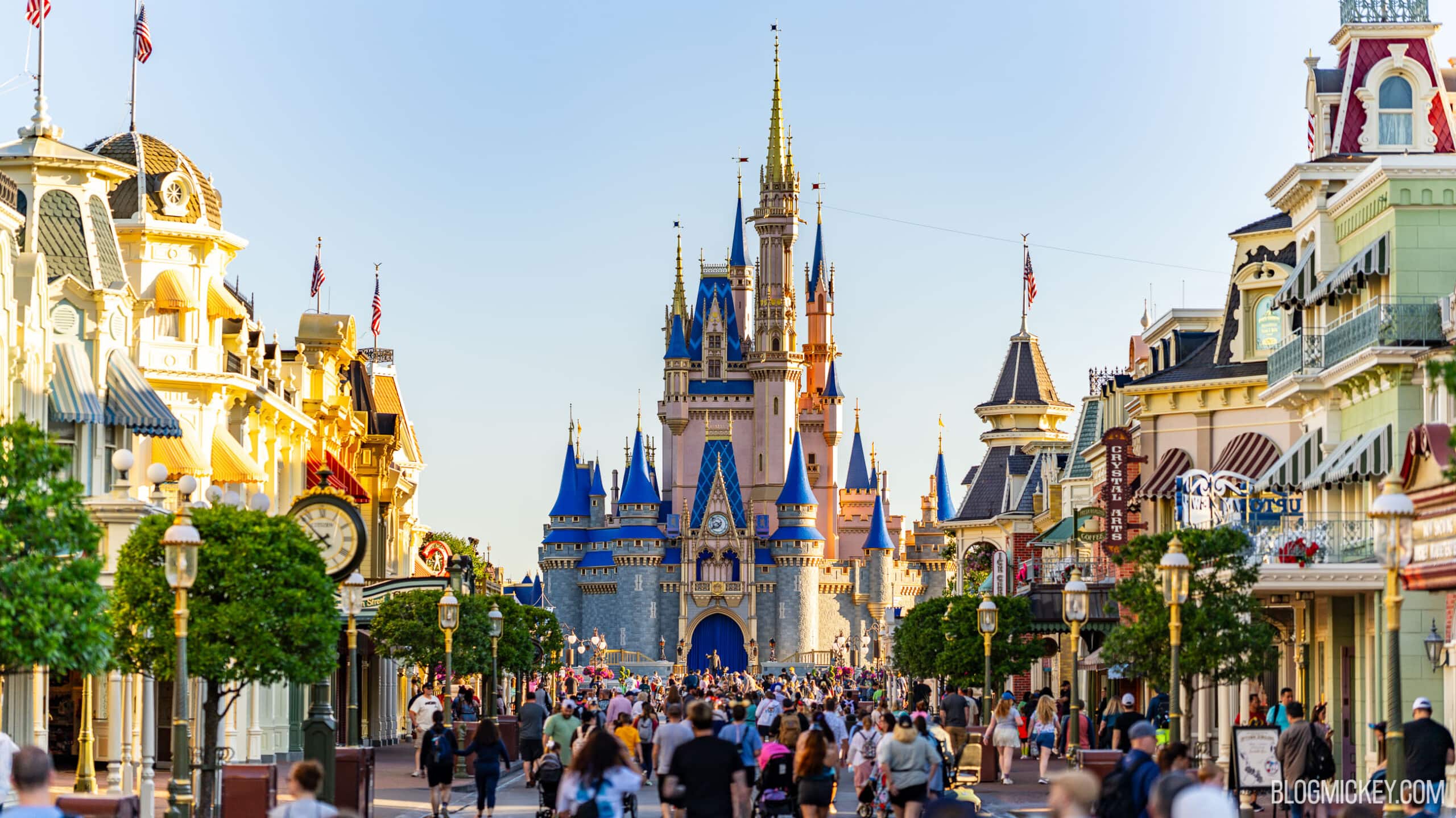 Main Street, U.S.A. at Magic Kingdom looking toward Cinderella Castle at golden hour, with crowds of guests walking down the street between the shops.