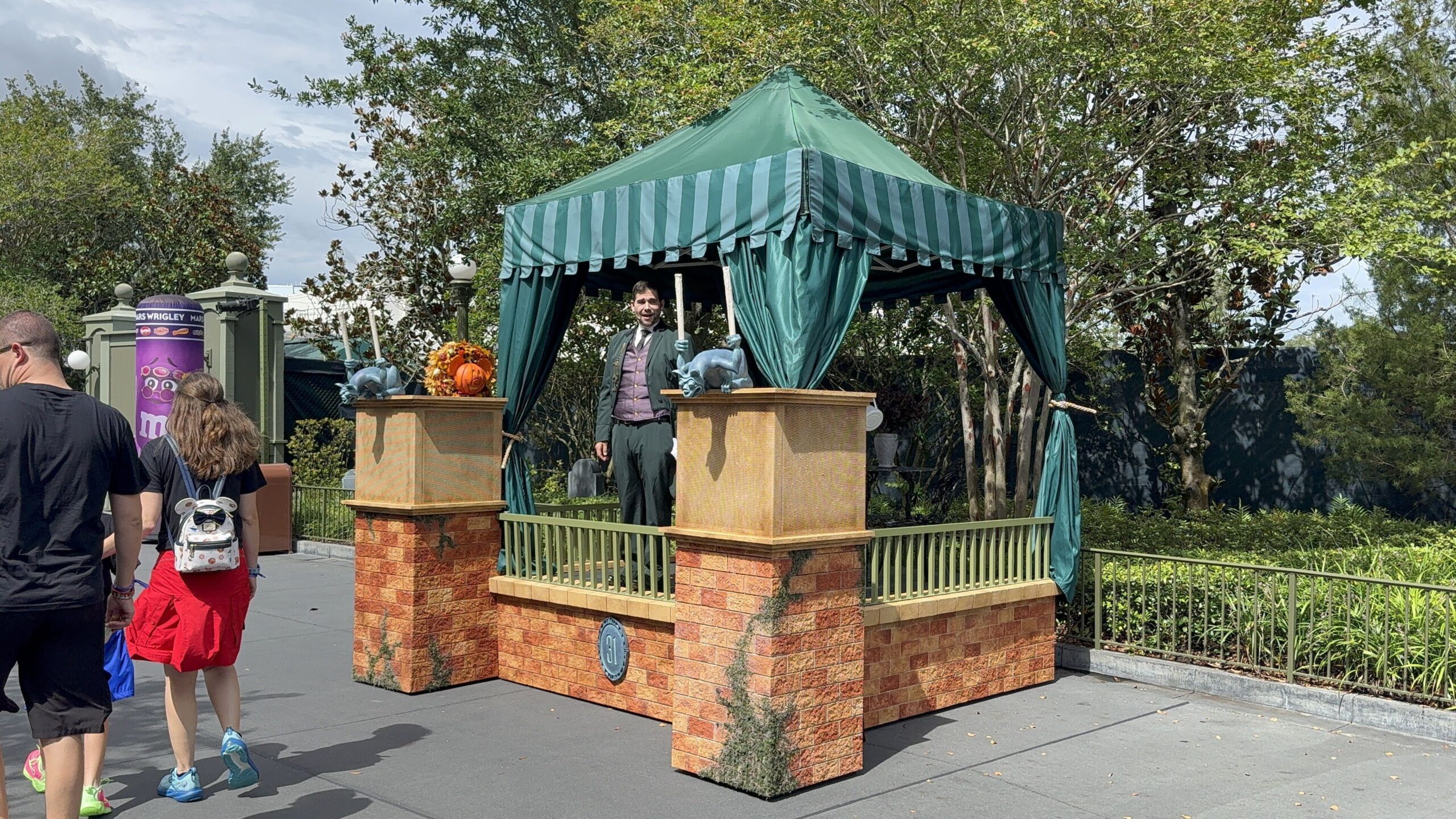 A man in a dark green suit and purple vest stands under a green striped canopy on a faux brick platform, with grey gargoyle statues on the corner posts, an autumn pumpkin arrangement, and a small plaque reading 31 on the brick base.