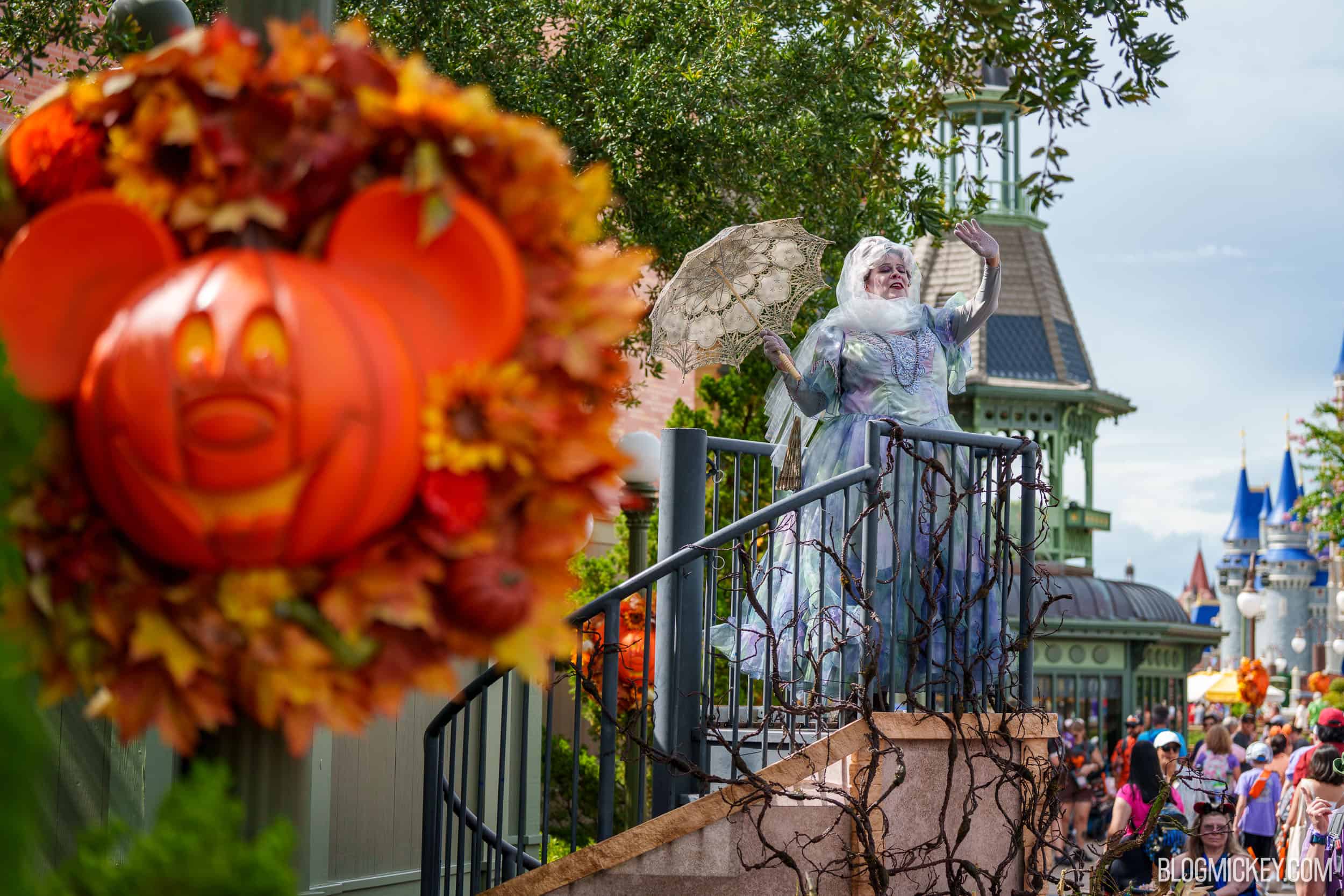 A pale ghostly woman in a shimmering blue and white gown waves from an elevated platform on Main Street, U.S.A., holding a lace parasol, with a Mickey pumpkin wreath in the foreground and Cinderella Castle behind her.
