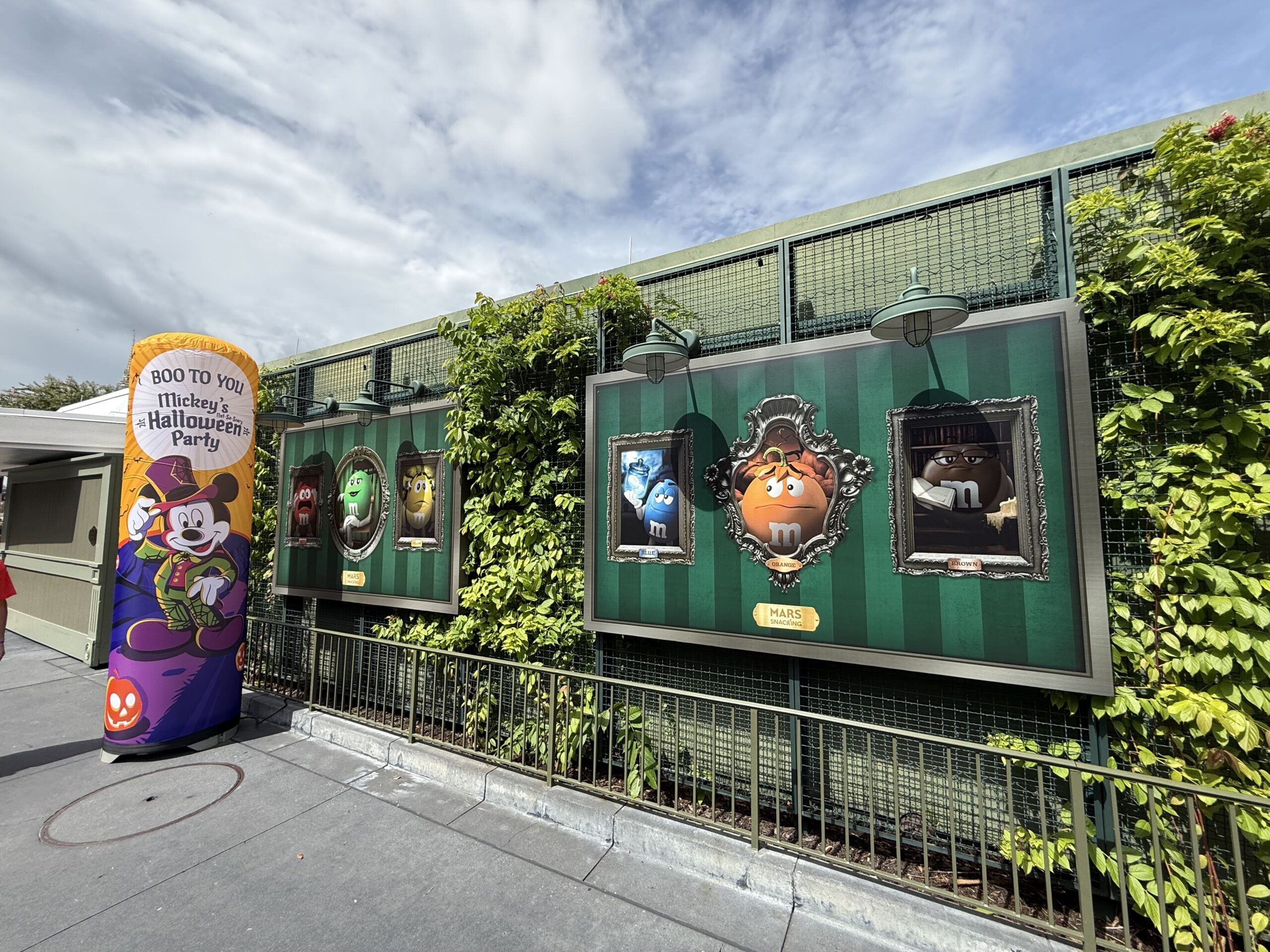 Two large display boards mounted on a green wall show M and M's candy characters inside ornate picture frames on green striped wallpaper, styled as Haunted Mansion portraits, next to an orange Boo To You Mickey's Not-So-Scary Halloween Party banner.