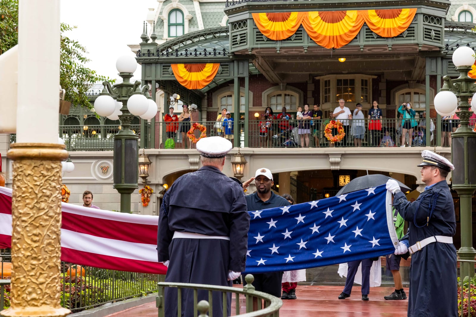 Honor guard members in dress uniform fold a large American flag in the rain at Magic Kingdom Town Square while guests watch from a balcony