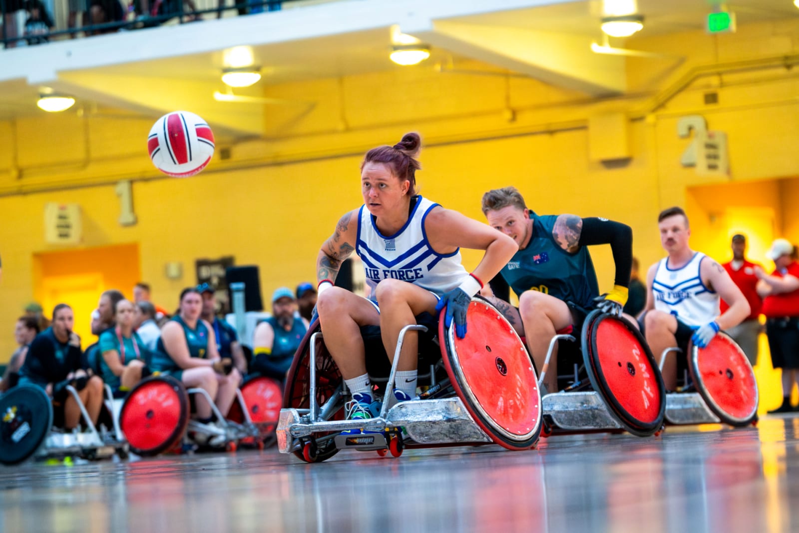 Athletes in Air Force jerseys and a green and gold Australian jersey chase a ball in racing wheelchairs on an indoor court