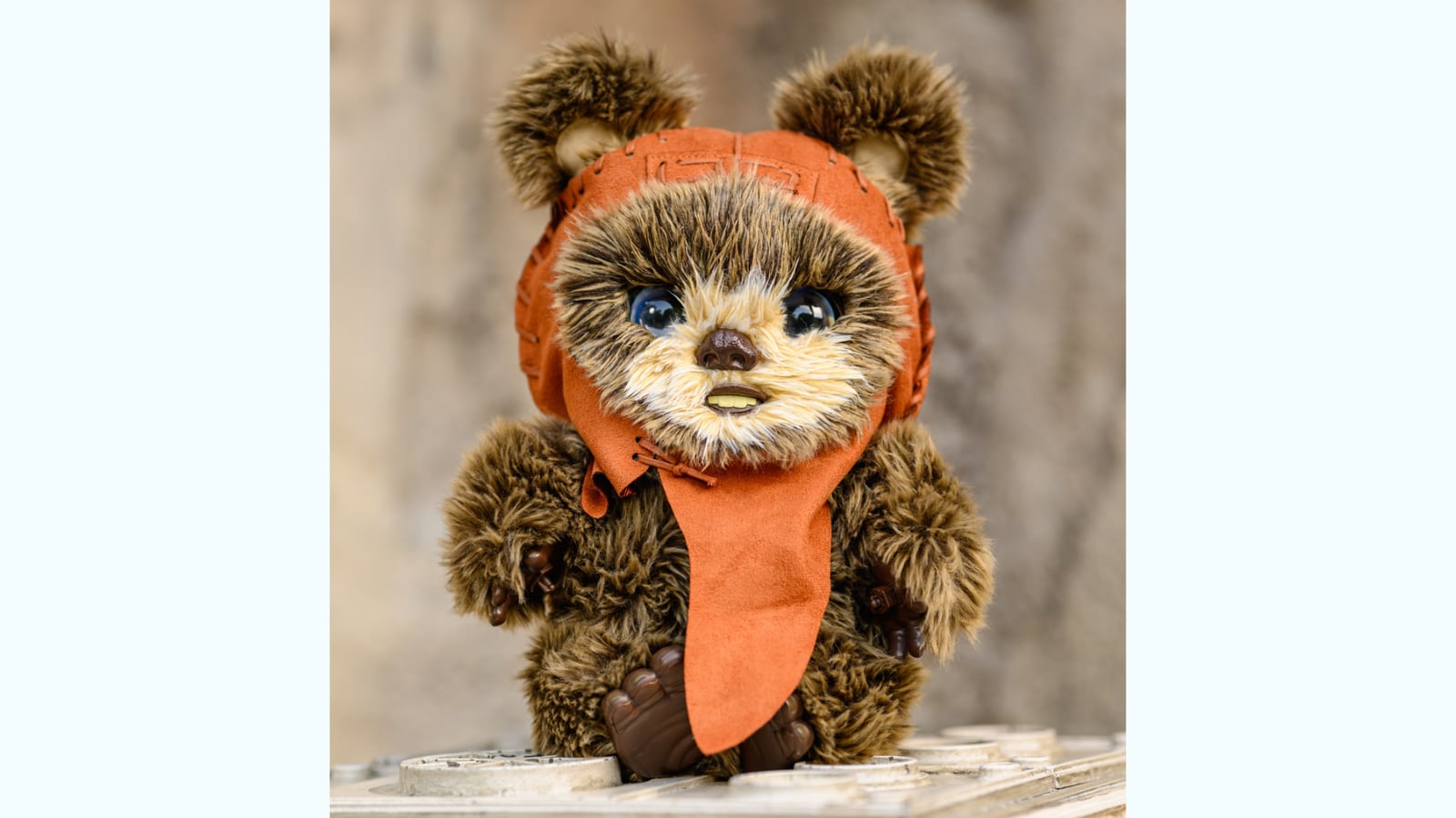 A small furry Ewok toy with large glossy blue eyes and an orange hood and cape stands on a weathered metal panel studded with round bolt heads, its brown plastic hands raised, against a blurred sandstone wall.