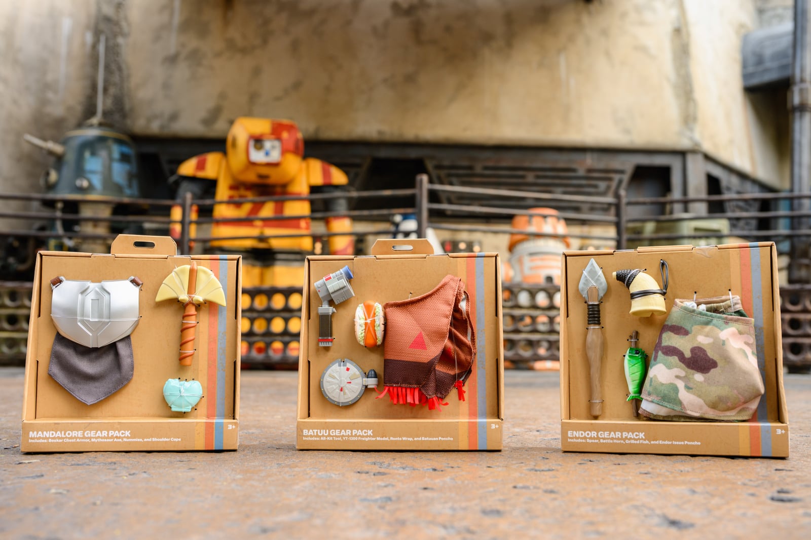 Three open cardboard retail boxes sit on a concrete floor in front of a blurred yellow droid. The left box is labeled Mandalore Gear Pack and holds silver chest armor with a grey shoulder cape below it, an axe and a small pale green item. The center box is labeled Batuu Gear Pack with a tool, a round freighter model, a wrapped food item and an orange poncho. The right box is labeled Endor Gear Pack with a spear, a battle horn, a green fish and a camouflage poncho.