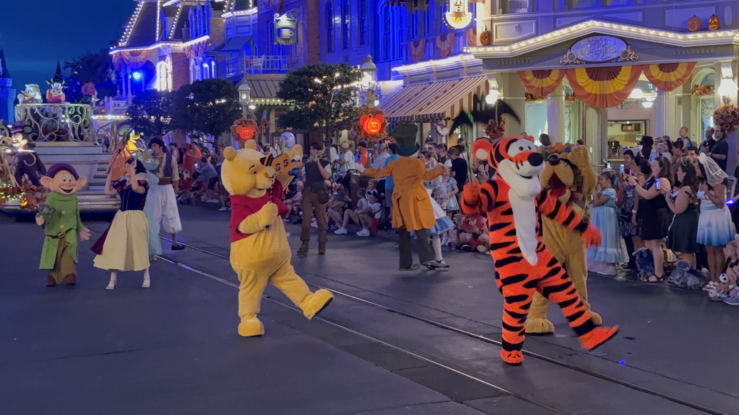 Winnie the Pooh and Tigger dancing down Main Street, U.S.A. at night during Mickey's Boo-To-You Halloween Parade, with the Mad Hatter dancing between the pair and Dopey and Snow White following farther back, guests lining both sides of the street, and orange Halloween bunting and Mickey pumpkin lights on the buildings.