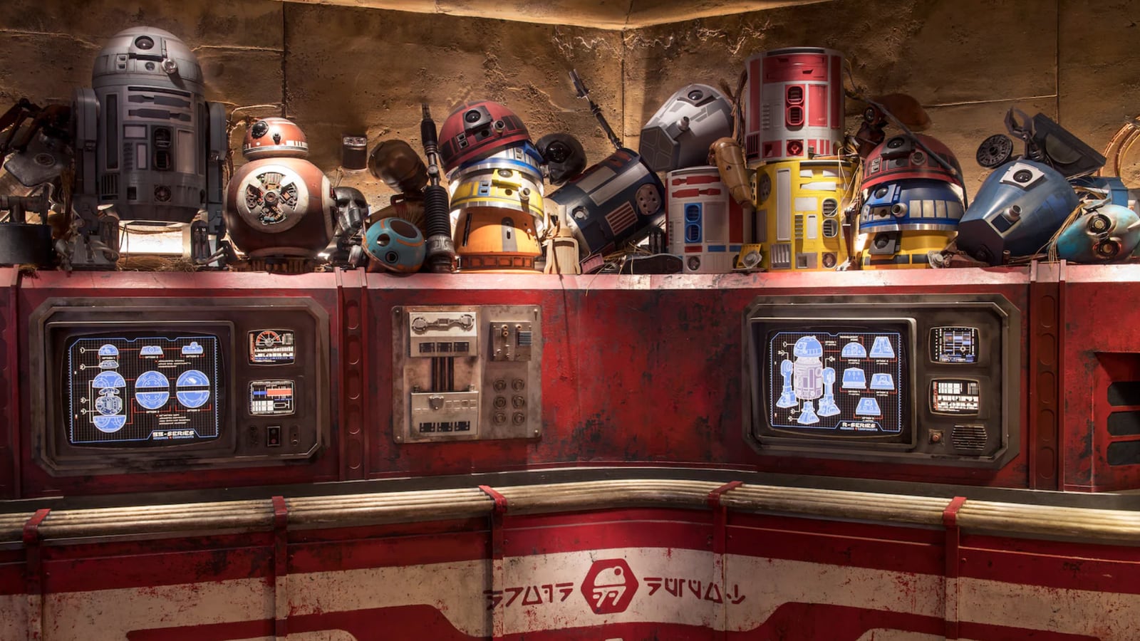 The interior of Droid Depot, with a crowded ledge of colorful droid domes, bodies and spare parts above a weathered red and white counter set with two lit blueprint screens showing BB-Series and R-Series droid components.