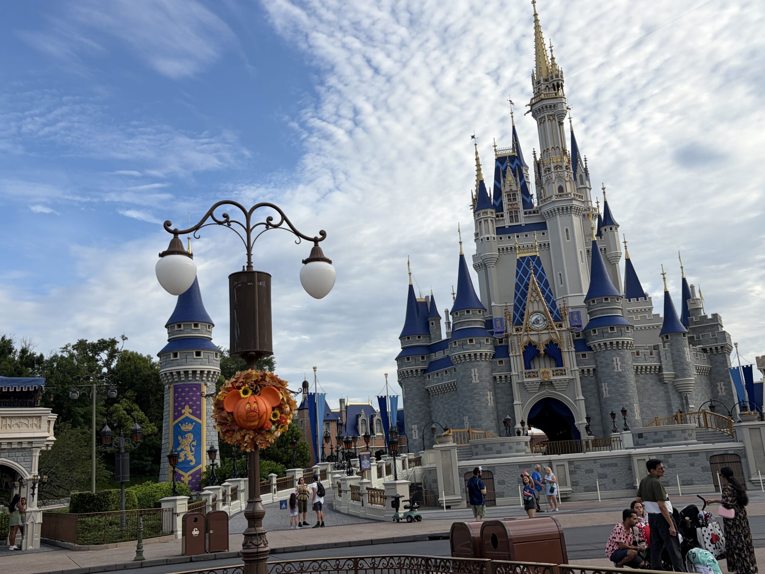Cinderella Castle in daylight under a partly cloudy sky, with a lamppost in the foreground carrying an autumn wreath built around a smiling Mickey-shaped jack-o'-lantern, and guests walking the pathways below.