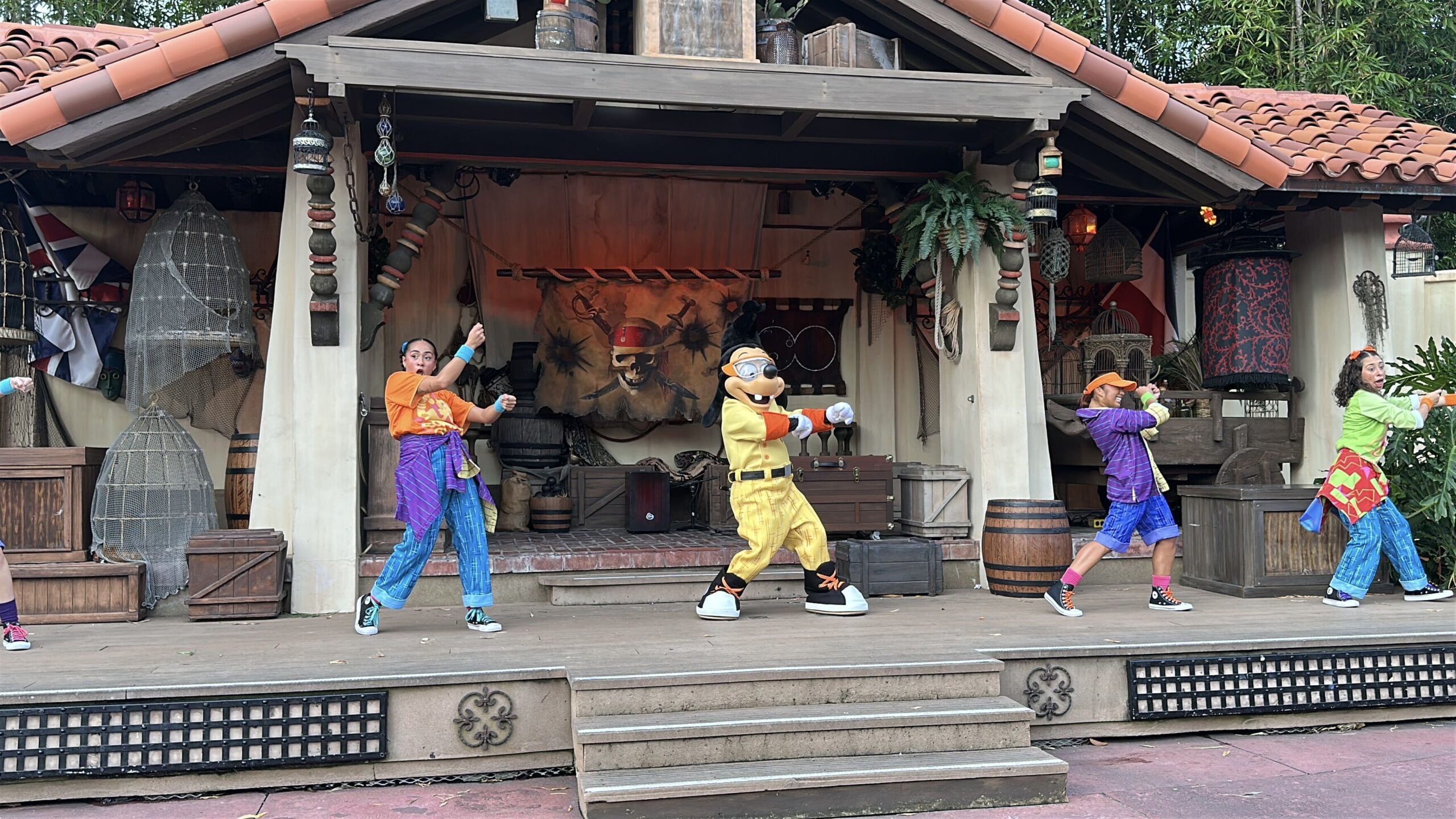Max Goof mid-dance-move in his yellow Powerline costume on the Adventureland stage, flanked by backup dancers in bright 1990s outfits in front of a painted pirate flag backdrop.