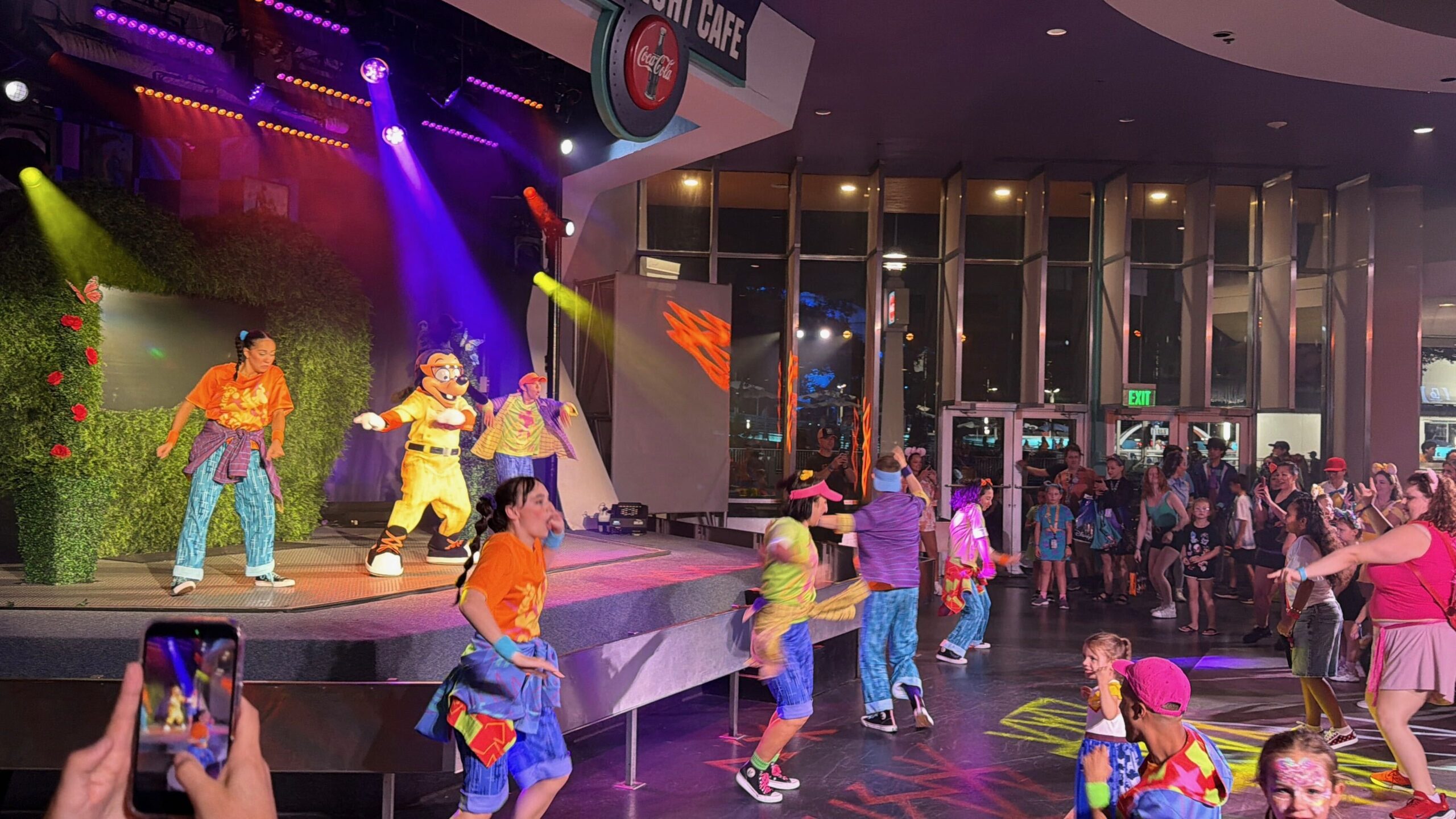 Night performance at Cosmic Ray's Starlight Cafe, with Max Goof and two dancers on a stage lit purple, pink and green, more dancers down on the pavement leading a crowd of guests in the routine.