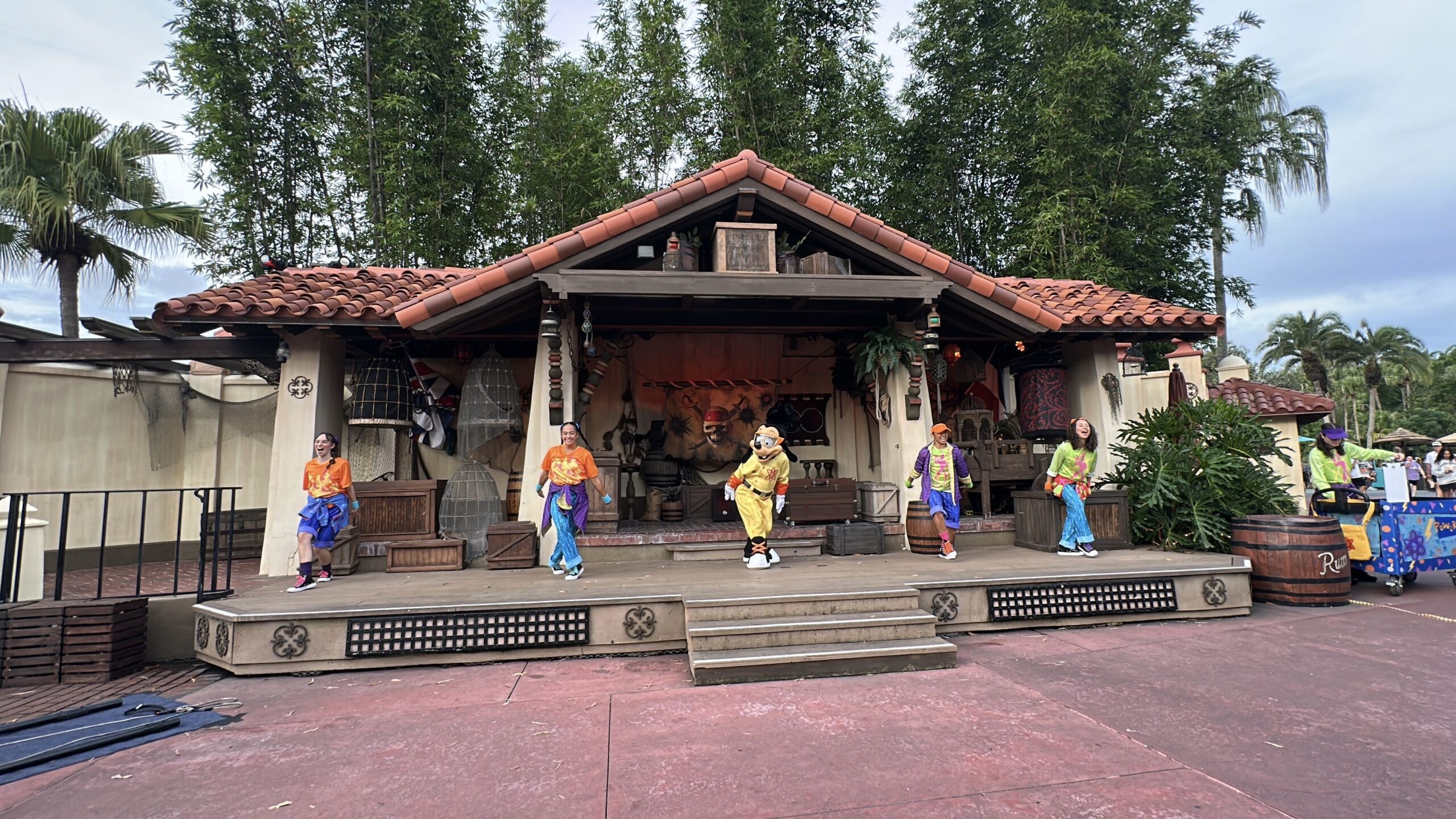 Max Goof in a yellow Powerline outfit dancing with four performers in bright 1990s clothes on the pirate-dressed Adventureland stage at Magic Kingdom, in daylight.