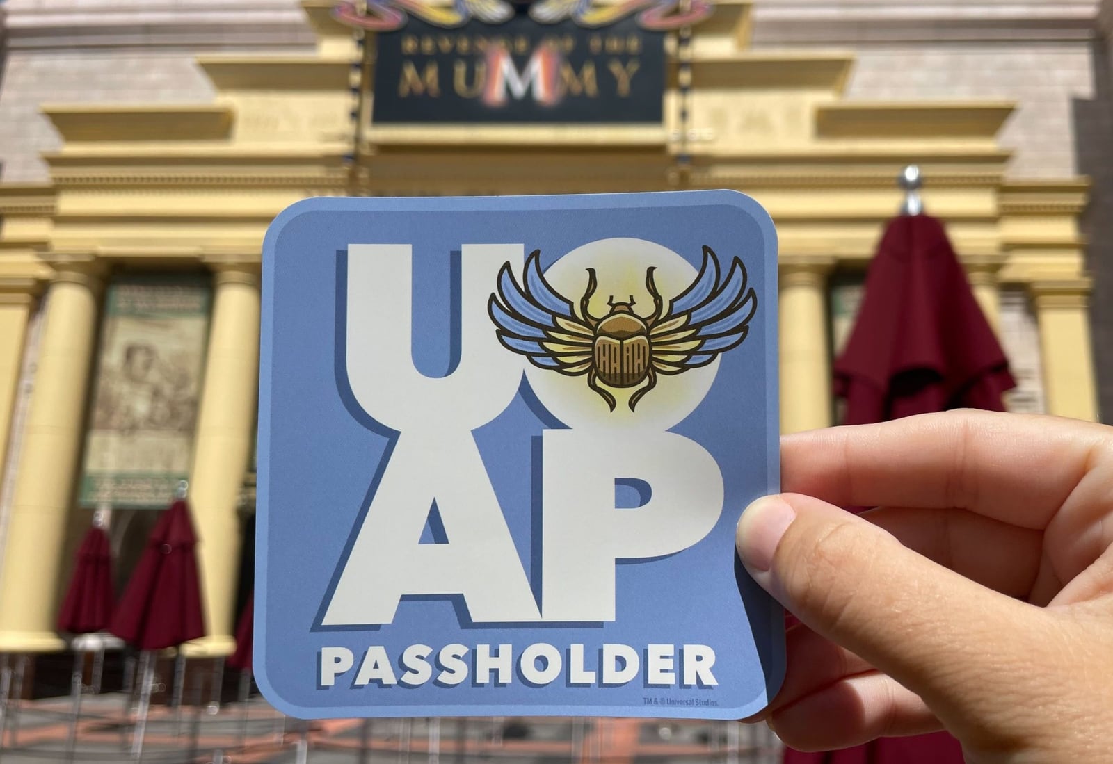 A hand holds up a square light blue magnet printed with a large white UOAP monogram, the word PASSHOLDER underneath, and a scarab beetle with a golden body and blue and cream wings. Behind it, out of focus, is the gold and stone Revenge of the Mummy attraction facade with its marquee and burgundy patio umbrellas.