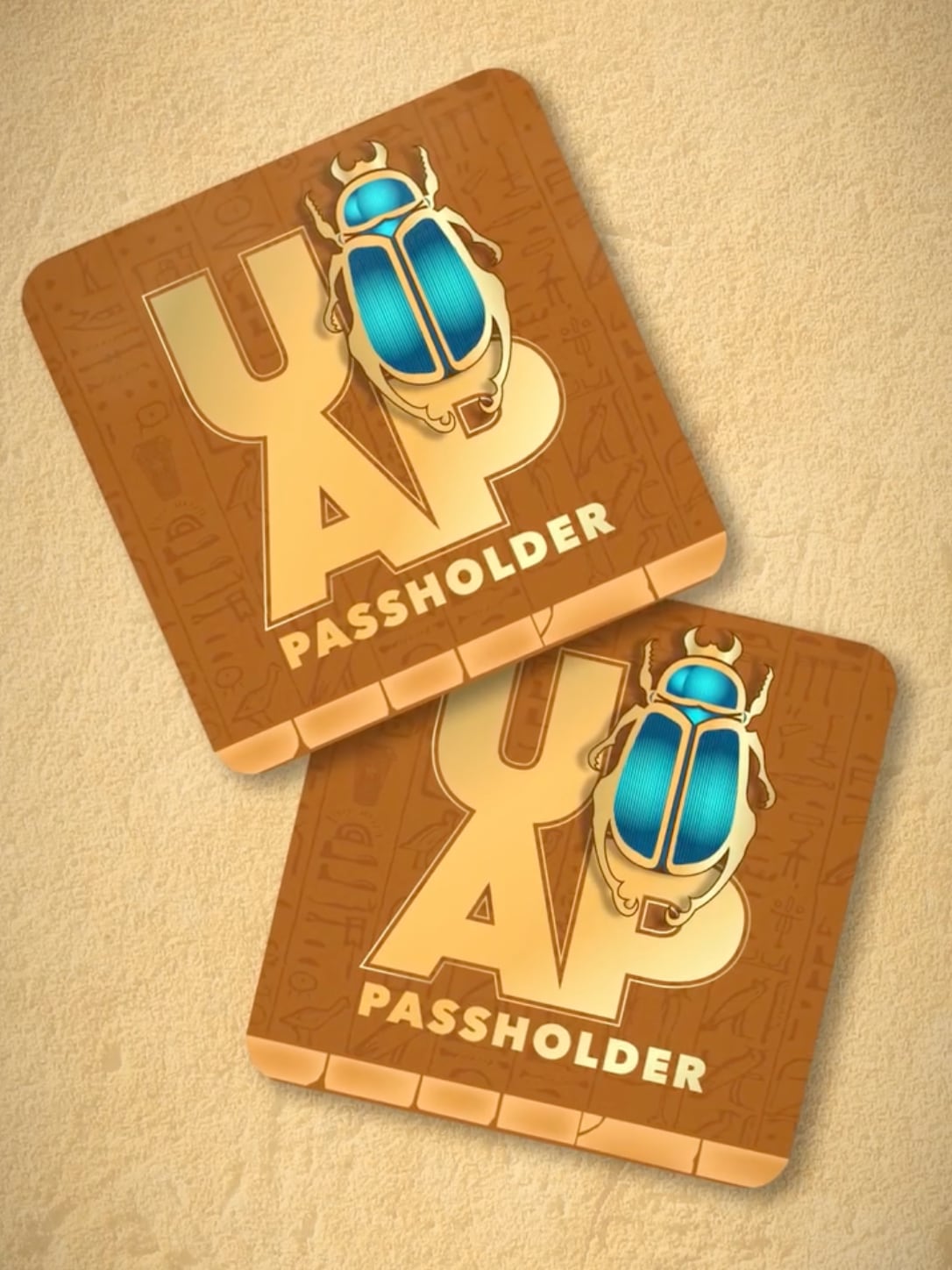 Two identical square magnets with rounded corners lie overlapping on a sandy background. Each has a warm brown surface carved with faint Egyptian hieroglyphs, a large gold UOAP monogram, the word PASSHOLDER along the bottom, and a metallic blue and gold scarab beetle filling the spot where the O belongs.