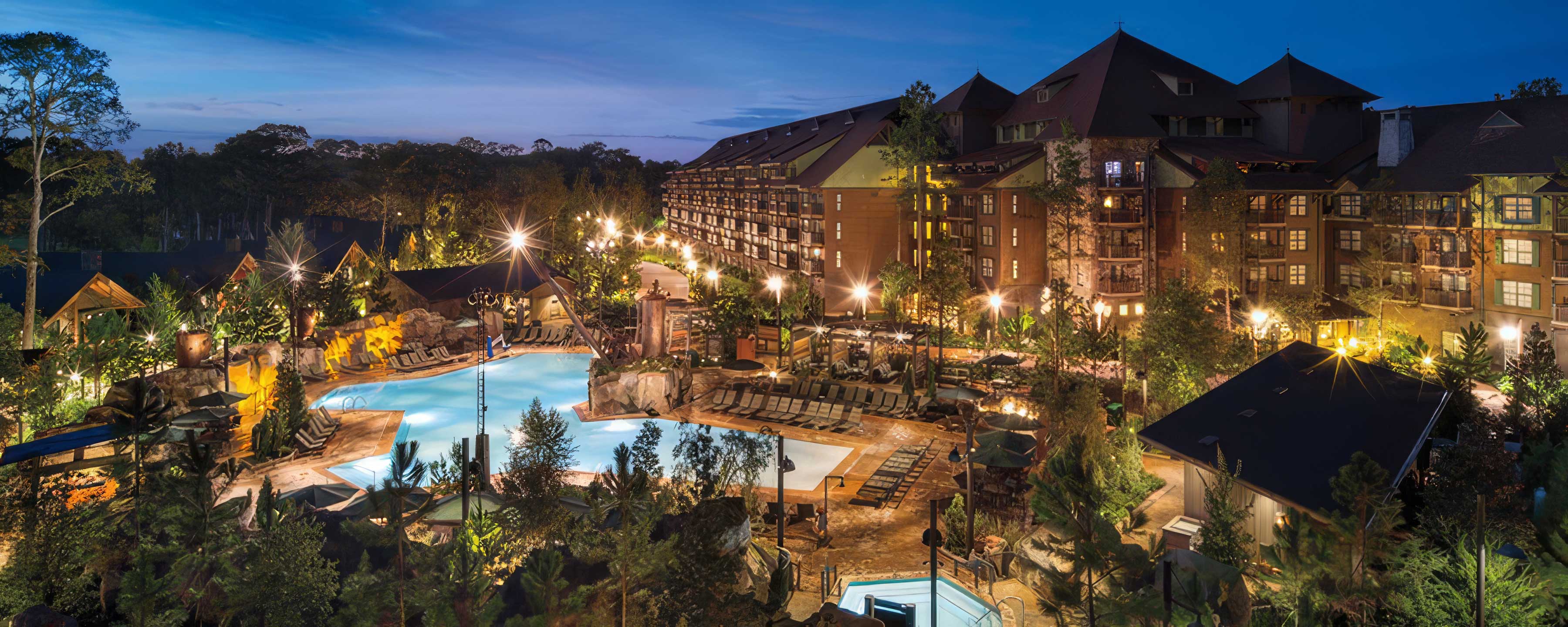 Disney's Wilderness Lodge at dusk, with lit guest balconies along a timber villa building above a themed pool area ringed by rockwork and pines.
