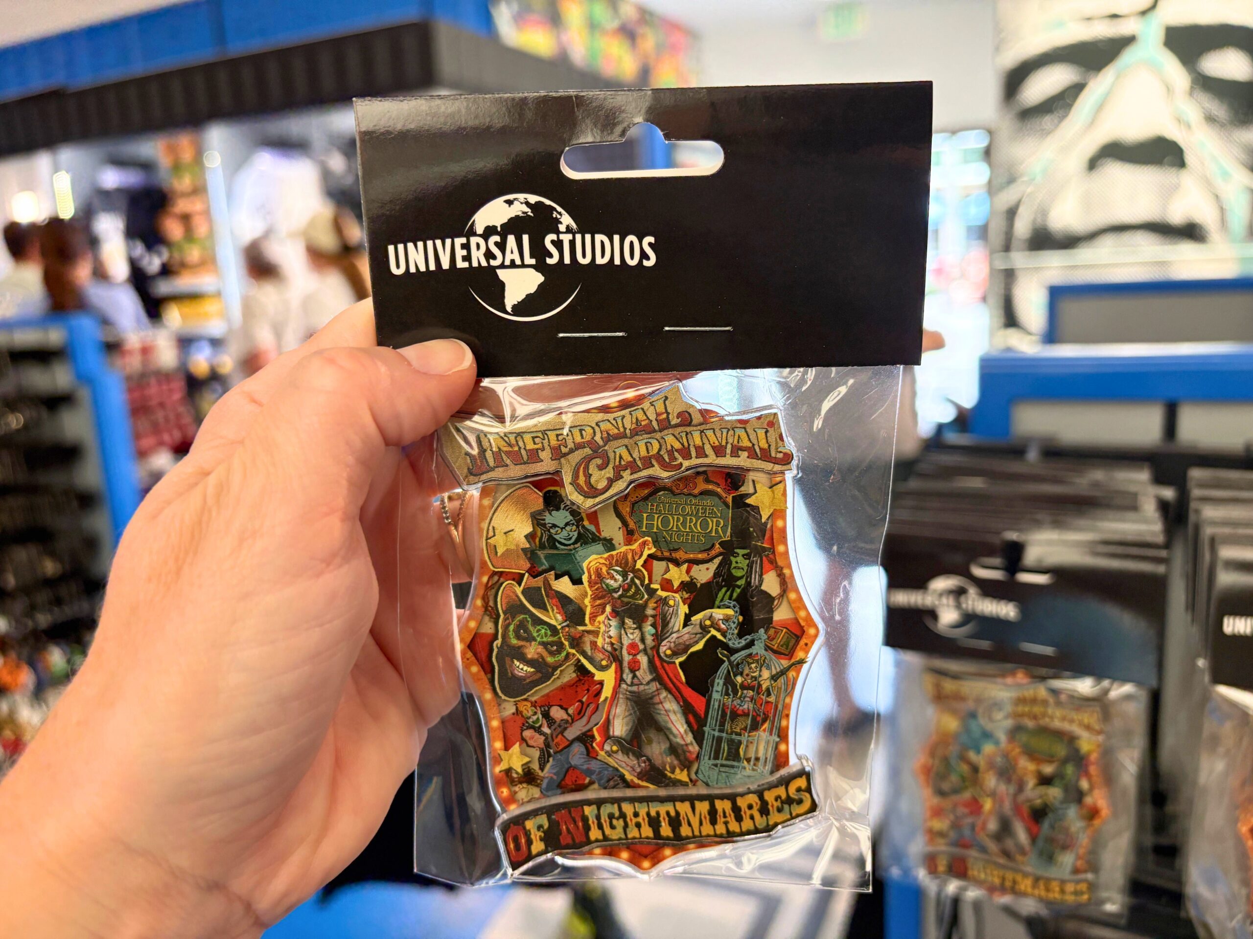 A guest holds up a packaged three-dimensional Infernal Carnival of Nightmares magnet on a black Universal Studios header card, showing the same carnival artwork with the Halloween Horror Nights logo near the top and OF NIGHTMARES lettering across the bottom.