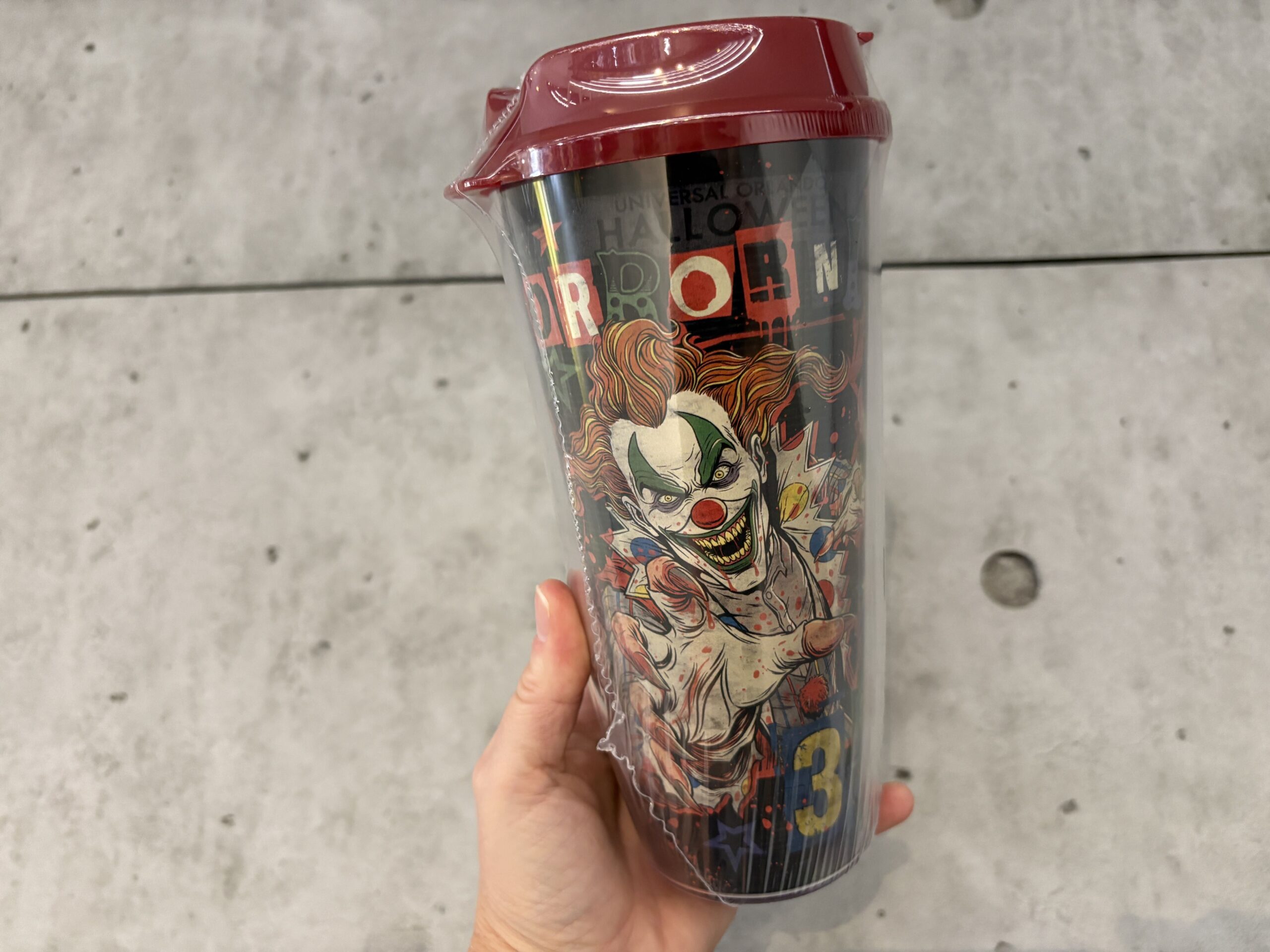 A guest holds the clear plastic refillable tumbler with a dark red lid. The wrap artwork shows Jack the Clown with orange hair and a wide toothy grin reaching out of the design with a bloodied hand, over black and red lettering.