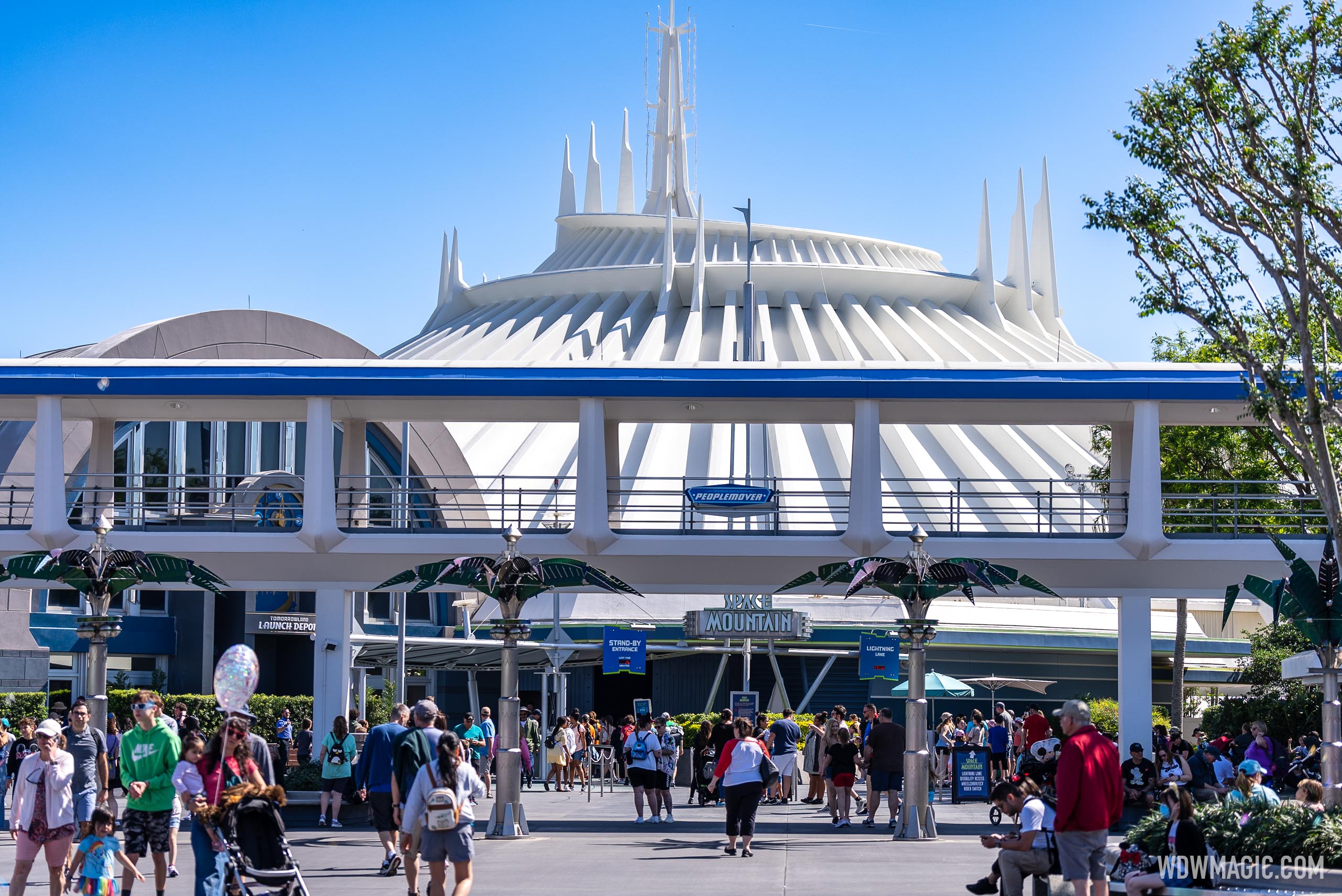The white ribbed cone of Space Mountain rising behind the Tomorrowland Transit Authority PeopleMover track, with the Space Mountain marquee, Stand-By Entrance and Lightning Lane banners and a crowd of guests below on a sunny day.